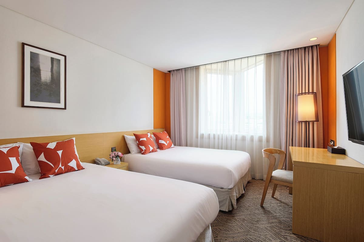 Une chambre d’hôtel avec deux lits, un bureau, une chaise, une lampe et une grande fenêtre avec des rideaux.