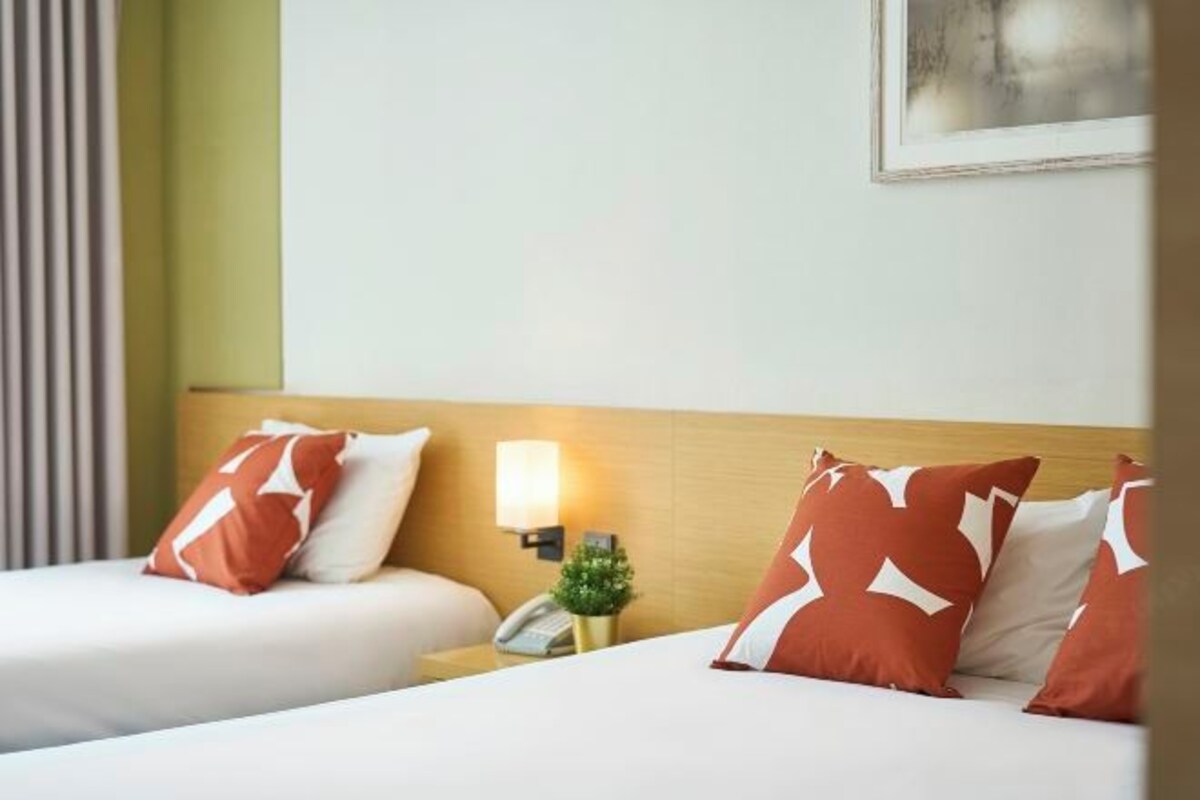 A hotel room with two beds, each with white and red patterned pillows, a bedside table with a lamp, and a small potted plant.