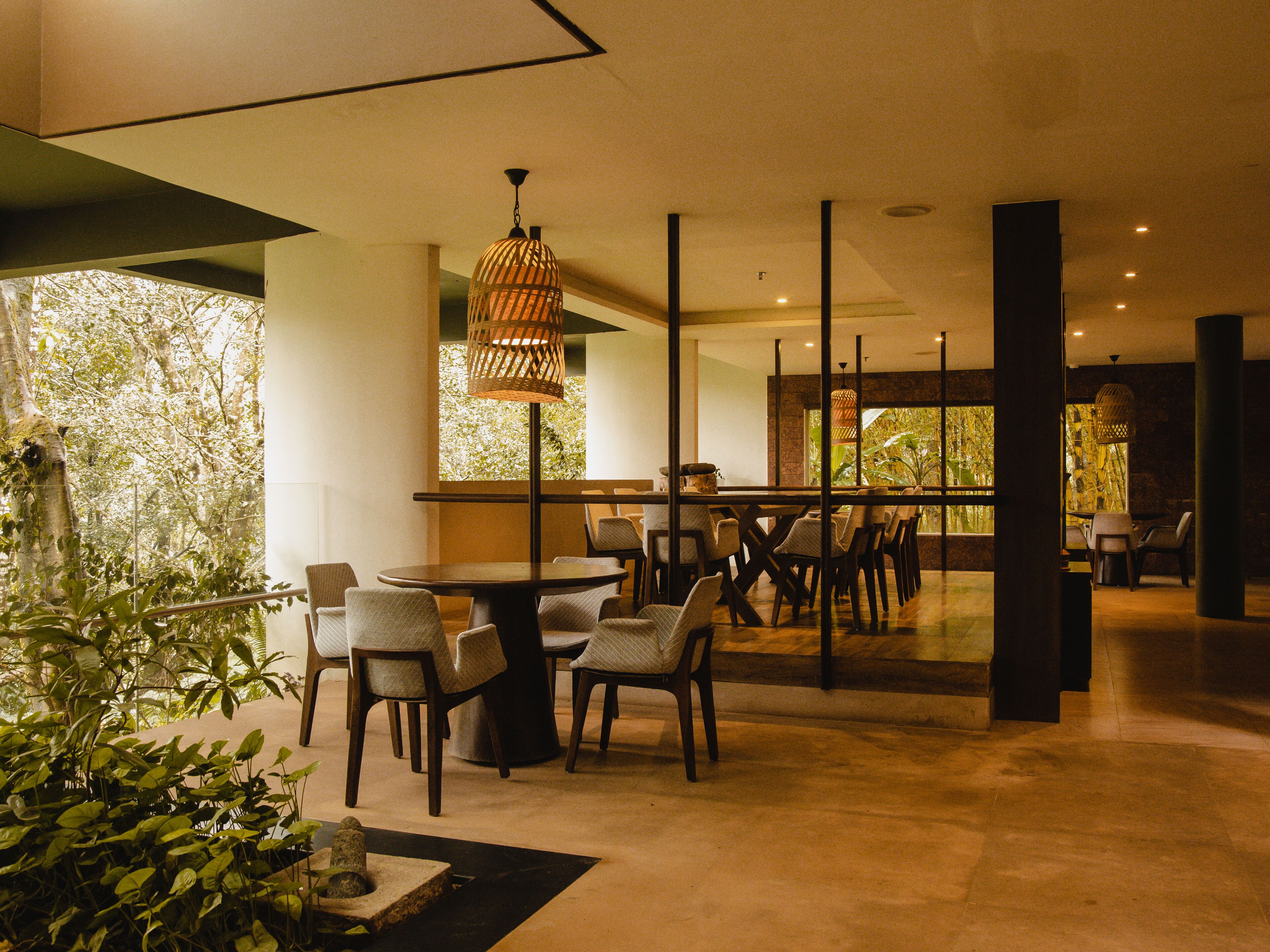A modern dining area with a round table and chairs, a hanging light fixture, and a view of greenery through large windows.
