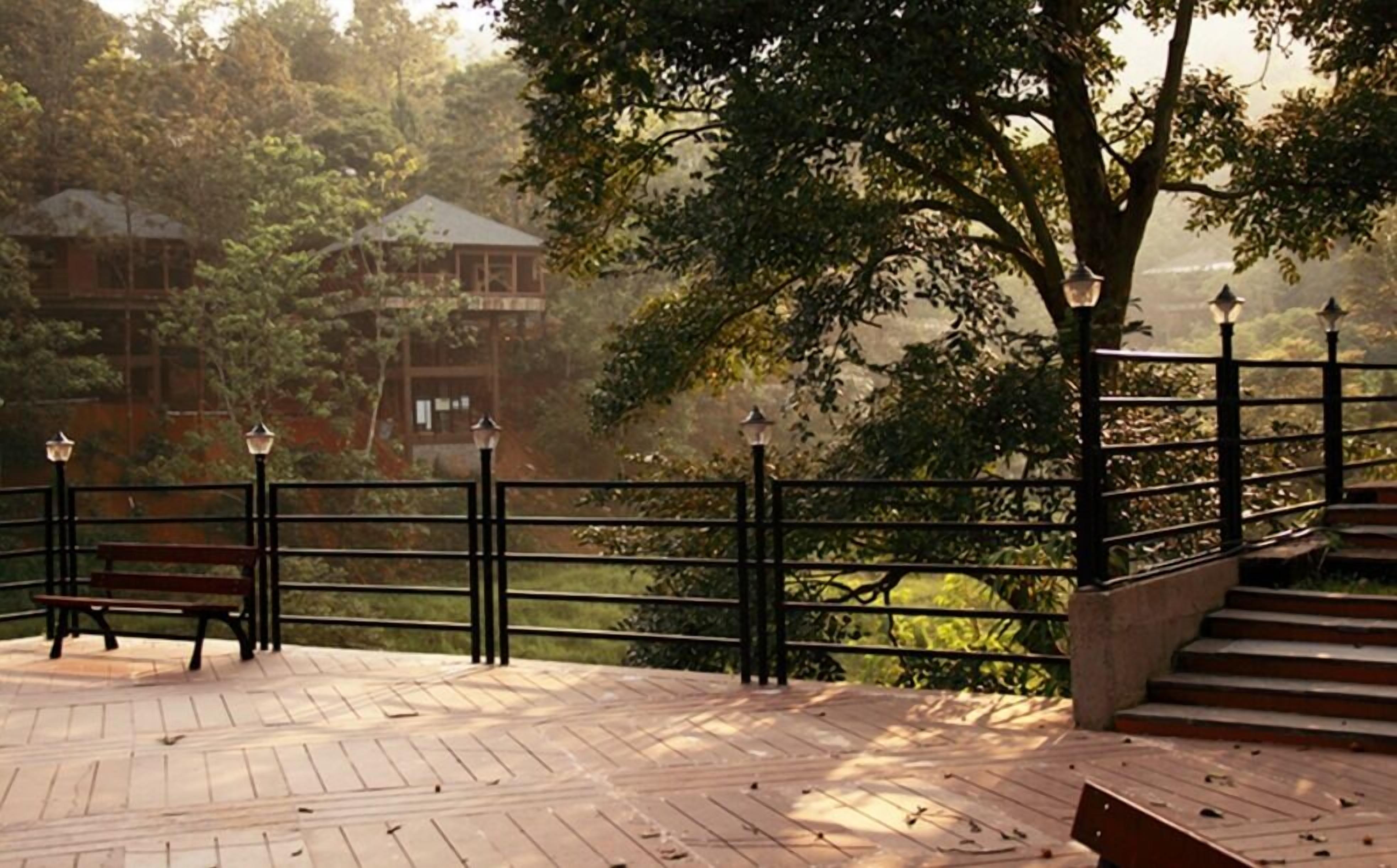 A wooden deck with a bench, a railing, and a view of a building surrounded by trees.