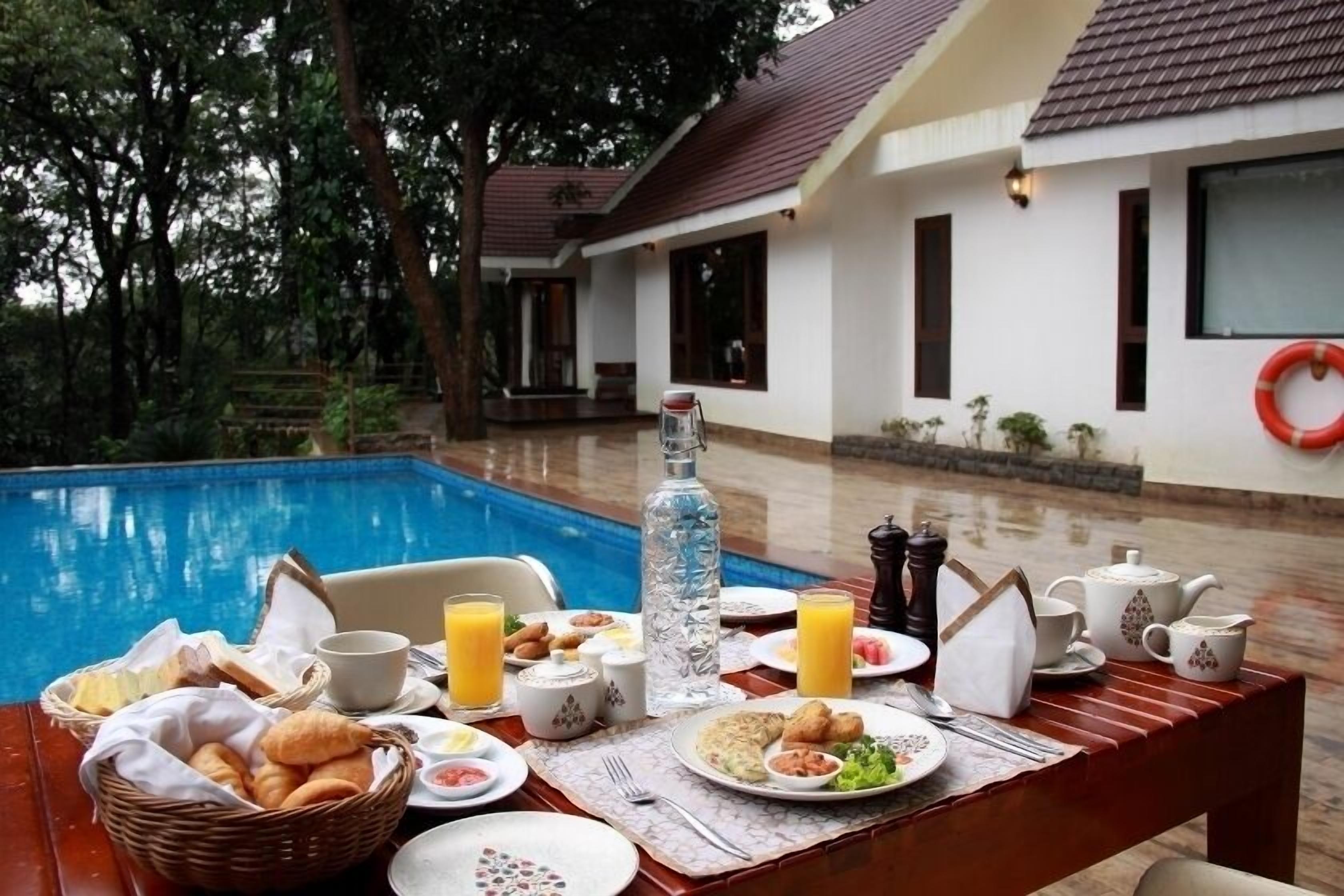 Mesa de desayuno con cruasanes, huevos y jugo junto a una piscina, con una casa y árboles al fondo.