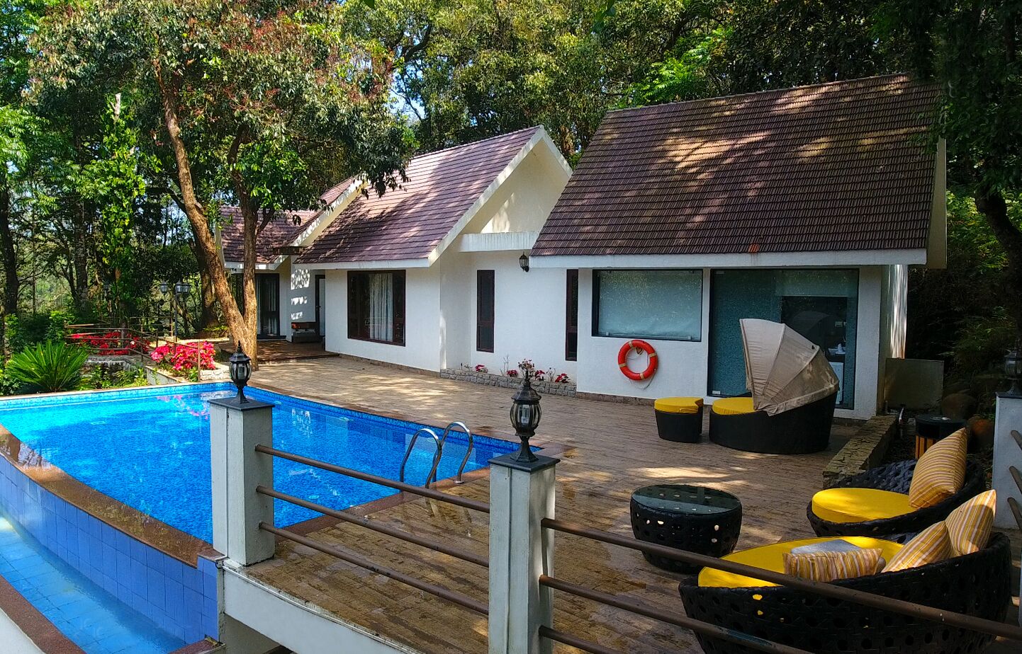 A swimming pool with a wooden deck, a covered seating area with cushions, and a white building with a tiled roof.