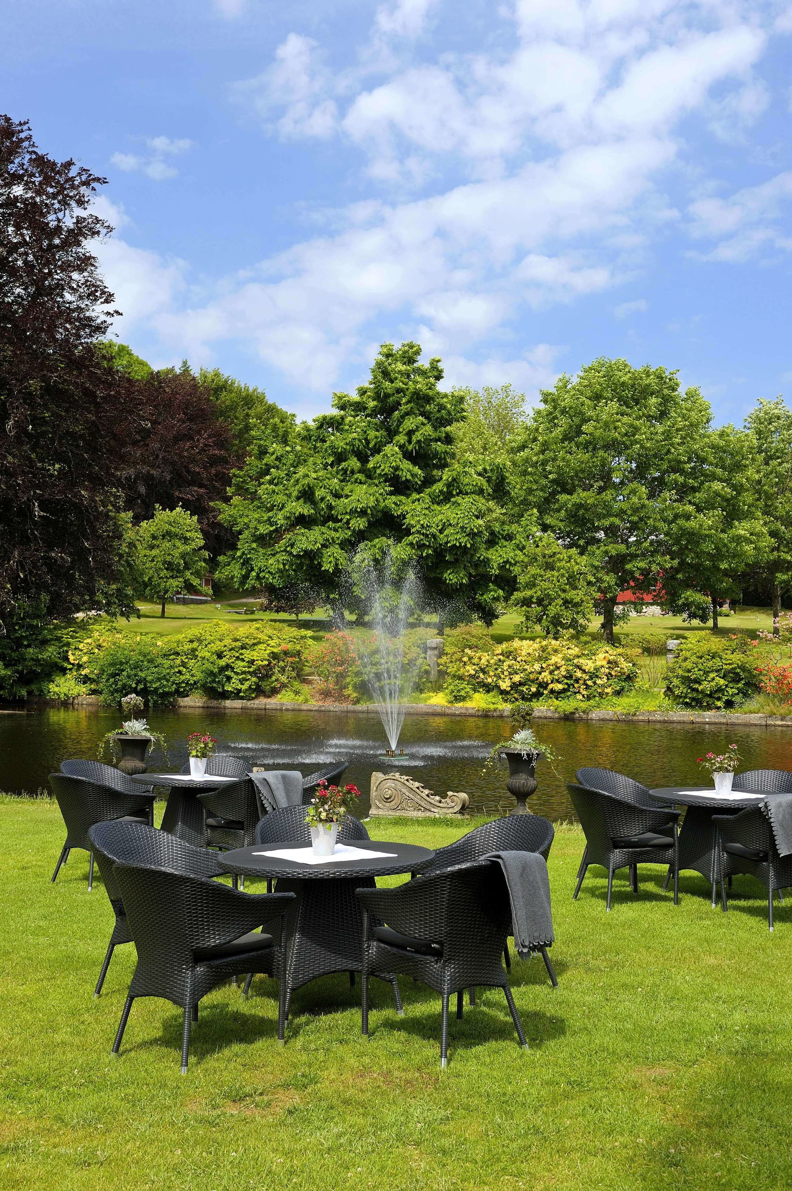 An outdoor seating area with wicker chairs and tables, a pond with a fountain, and lush greenery in the background.
