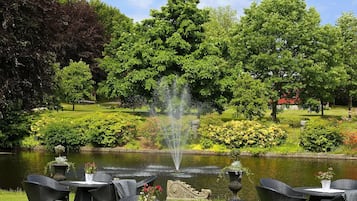 An outdoor seating area with wicker chairs and tables, a pond with a fountain, and lush greenery in the background.