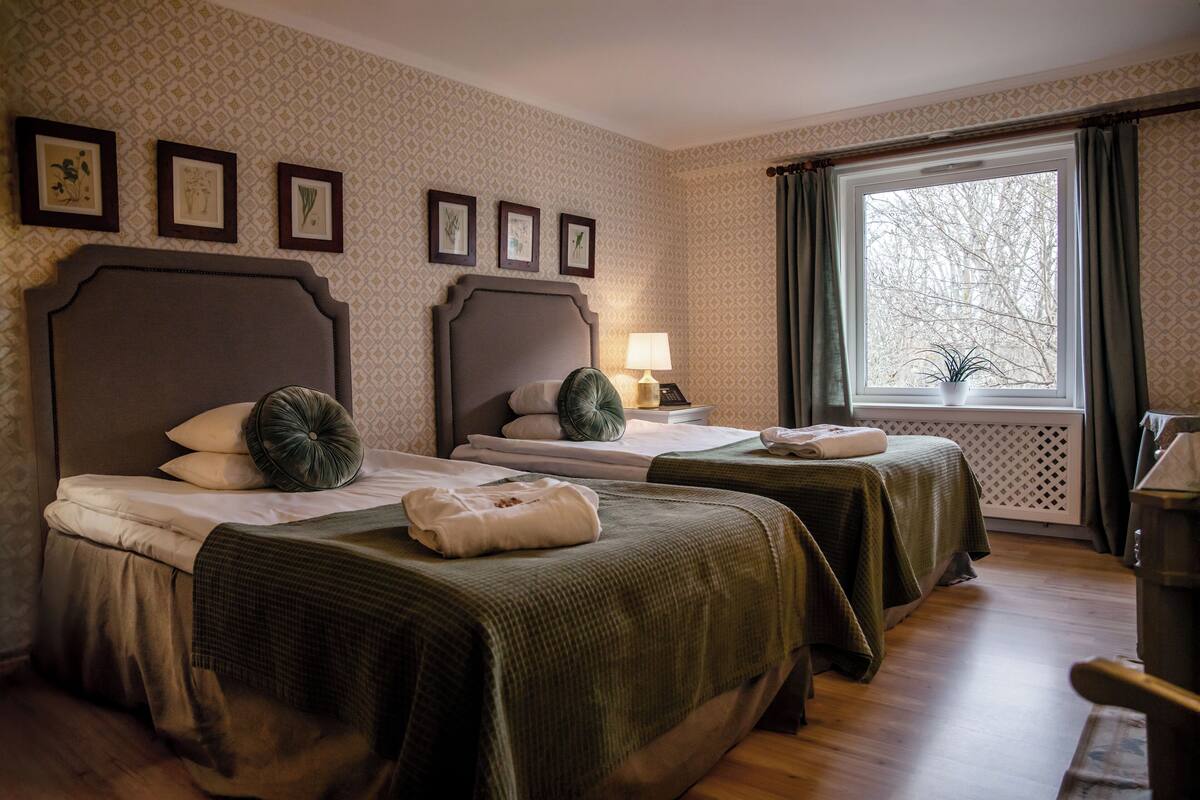 A hotel room with two beds, a window with curtains, a lamp, and framed pictures on the wall.