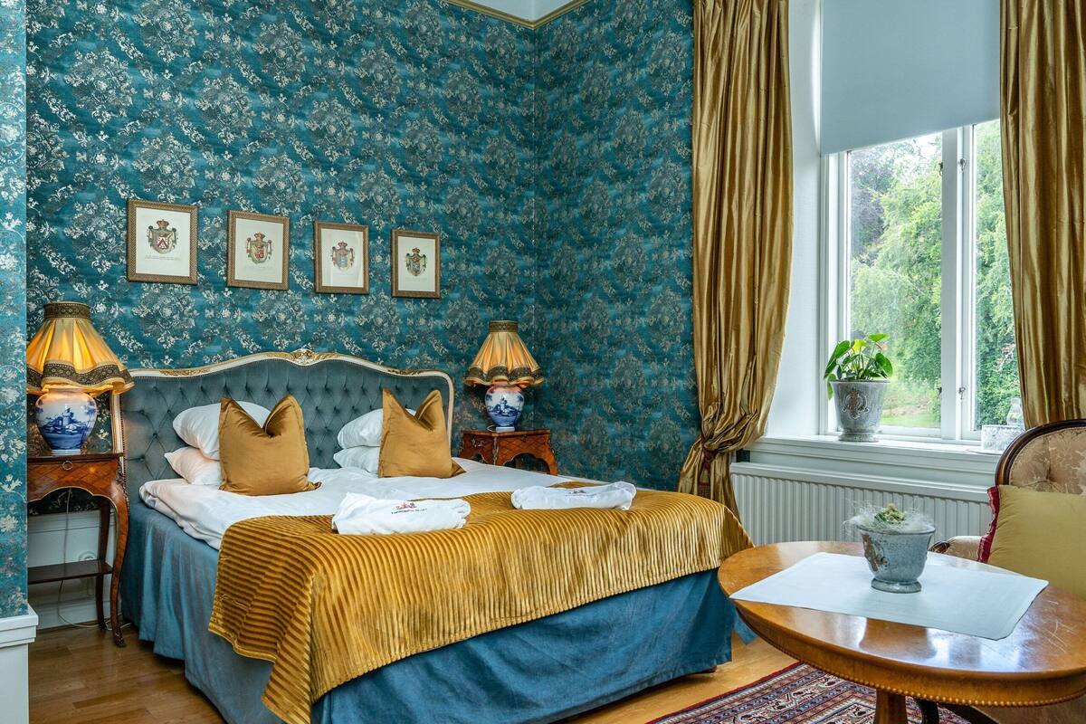 A bedroom with a large bed, two bedside tables with lamps, a window with curtains, and a view of greenery outside.