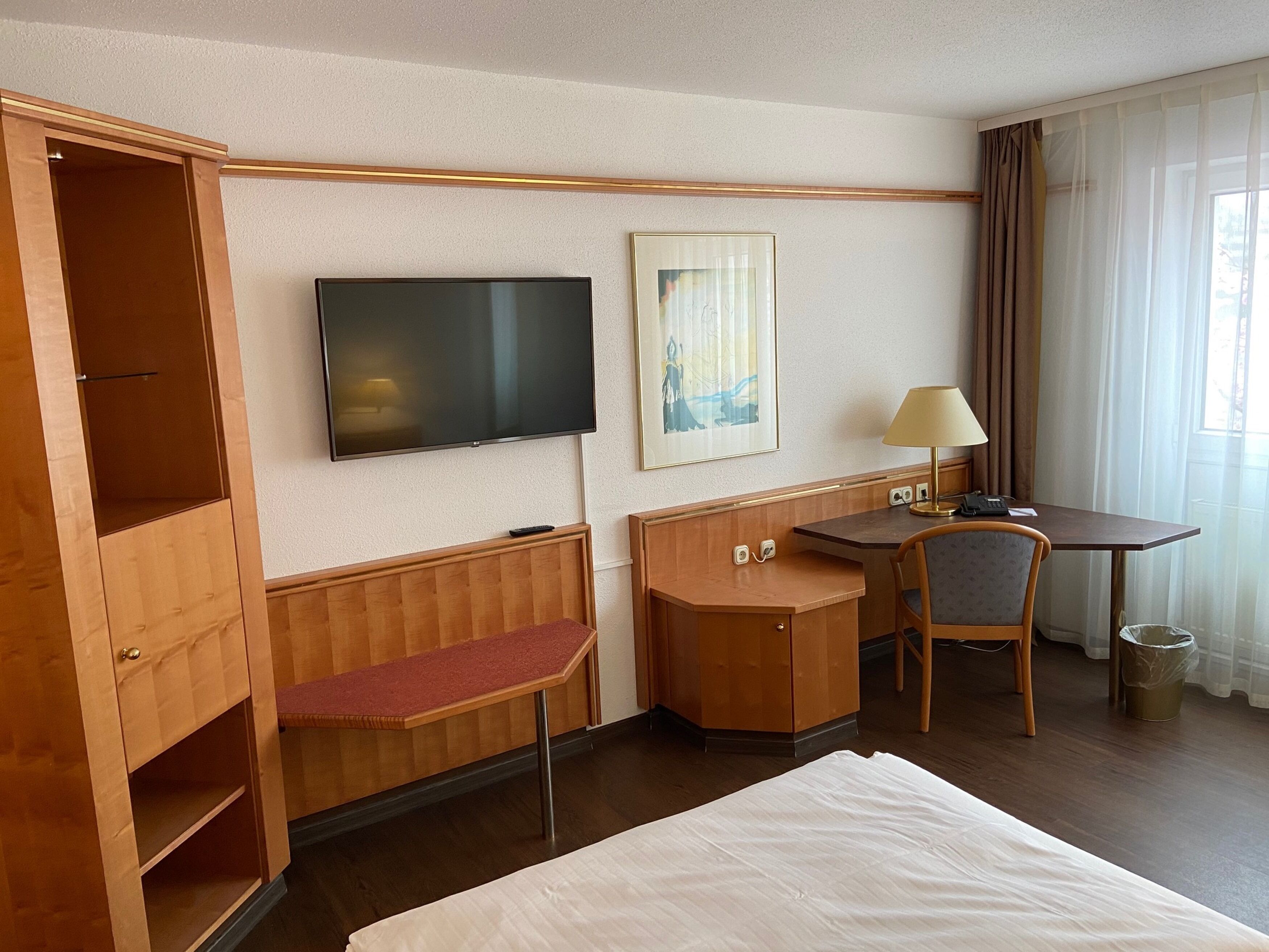 Ein Hotelzimmer mit Bett, Schreibtisch, Sessel, Fernseher, einem Gemälde und einem Kleiderschrank.