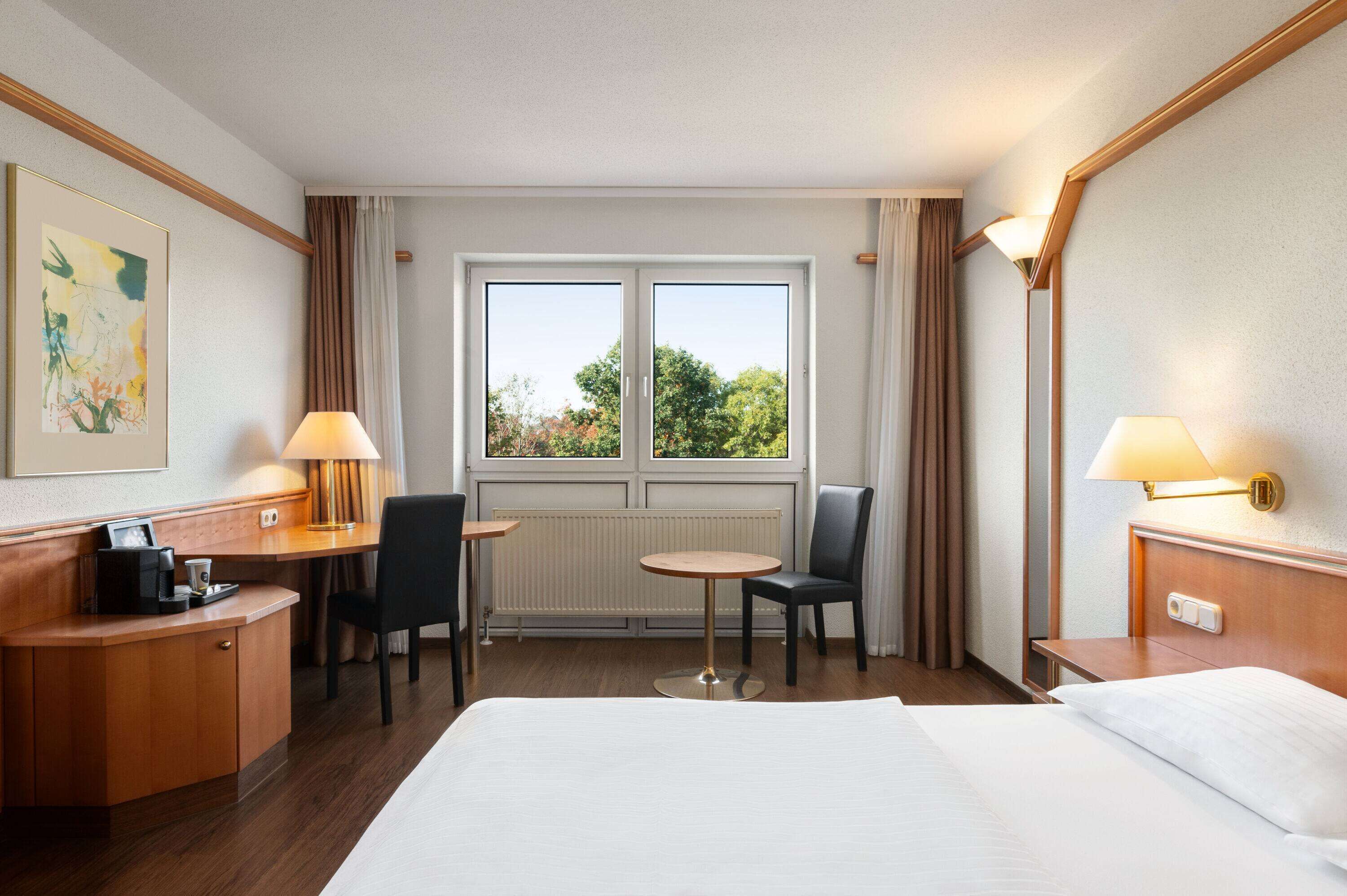 A hotel room with a bed, a desk, two chairs, a small table, and a window with curtains.