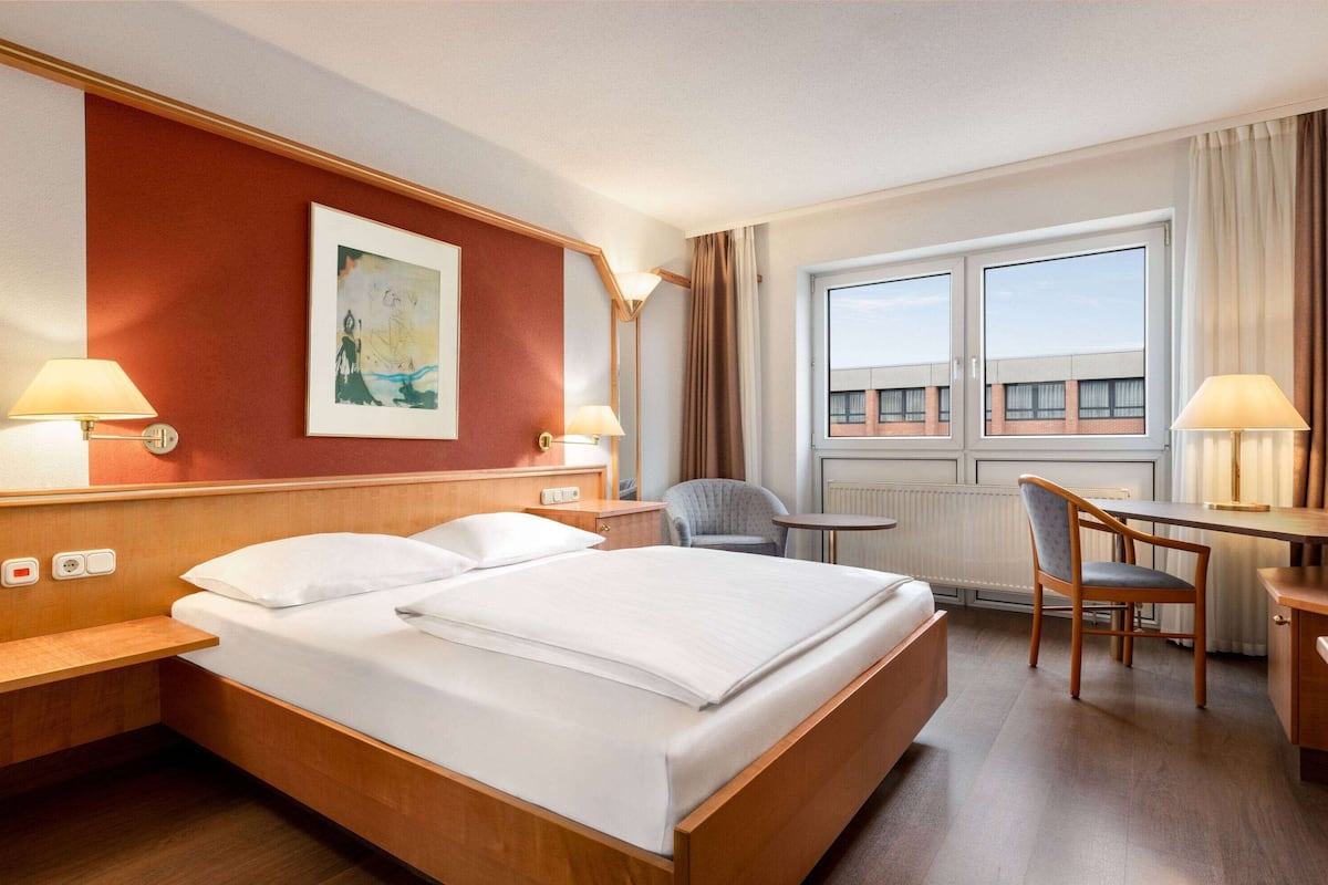 Ein Hotelzimmer mit einem großen Bett, einem Schreibtisch, einem Sessel und einem Fenster mit Blick auf Gebäude.