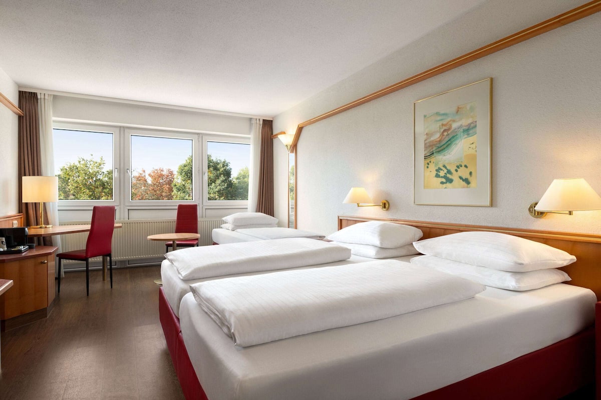 Ein Hotelzimmer mit einem großen Bett, einem Schreibtisch mit Stuhl und einem Fenster mit Blick auf Bäume.