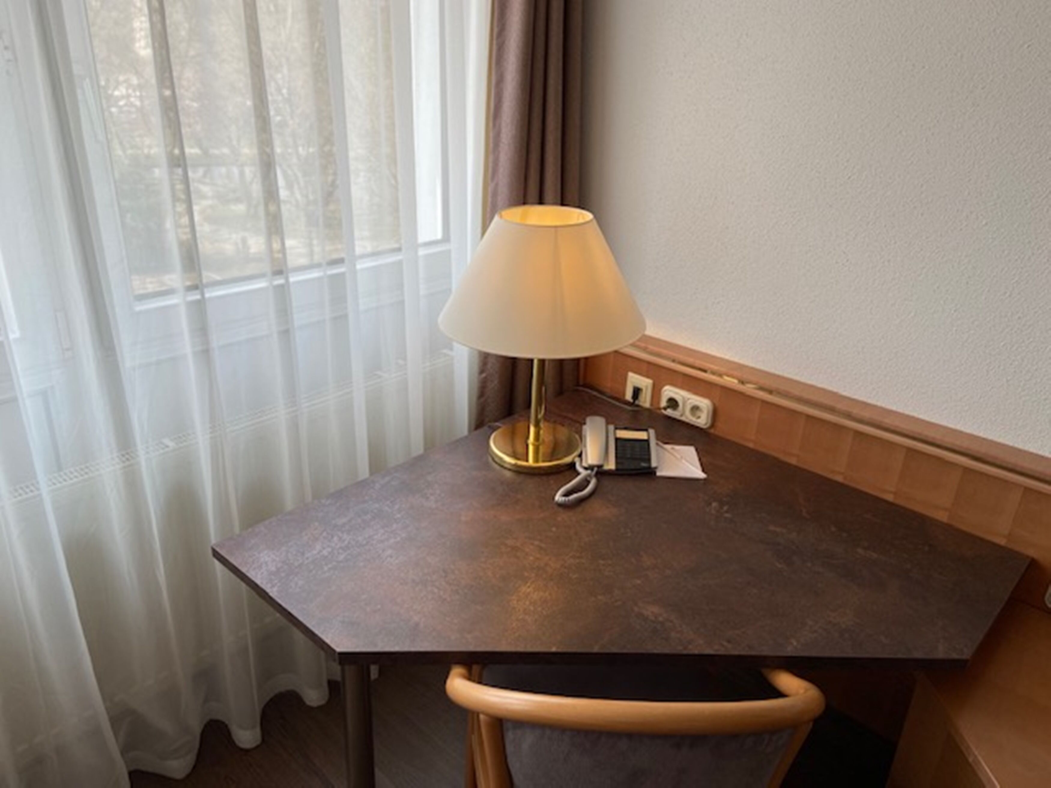 A small, dark wooden desk with a lamp, a telephone, and a notepad in a hotel room.