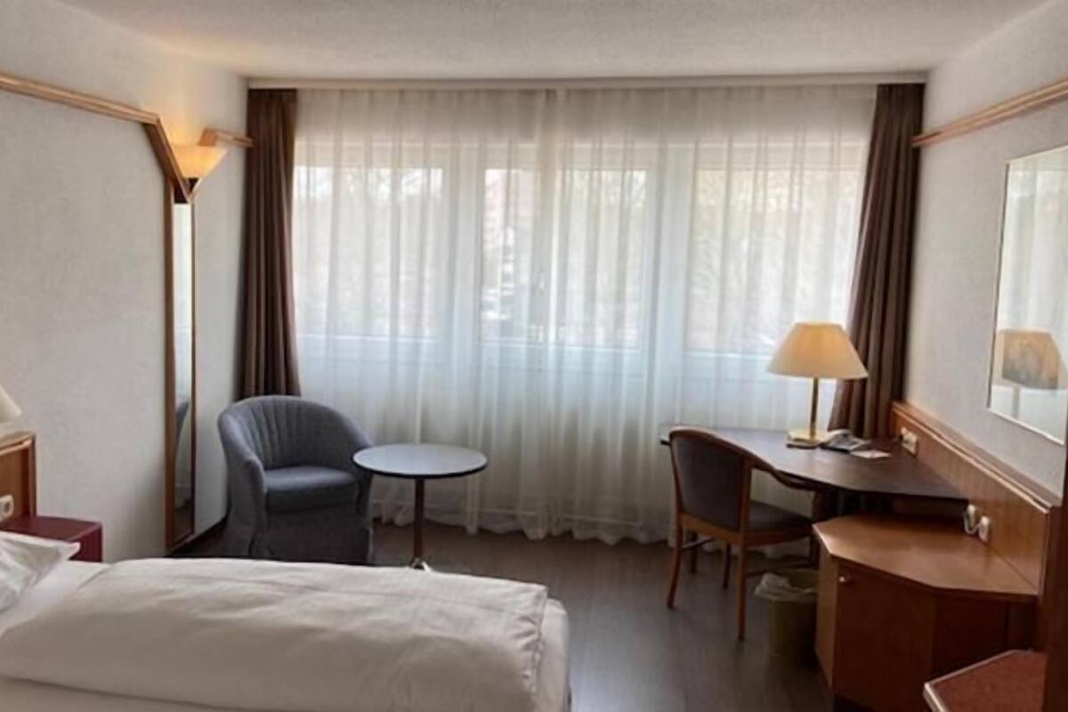 Ein Hotelzimmer mit Bett, Schreibtisch, Stuhl, kleinem Tisch und einem Fenster mit Vorhängen.
