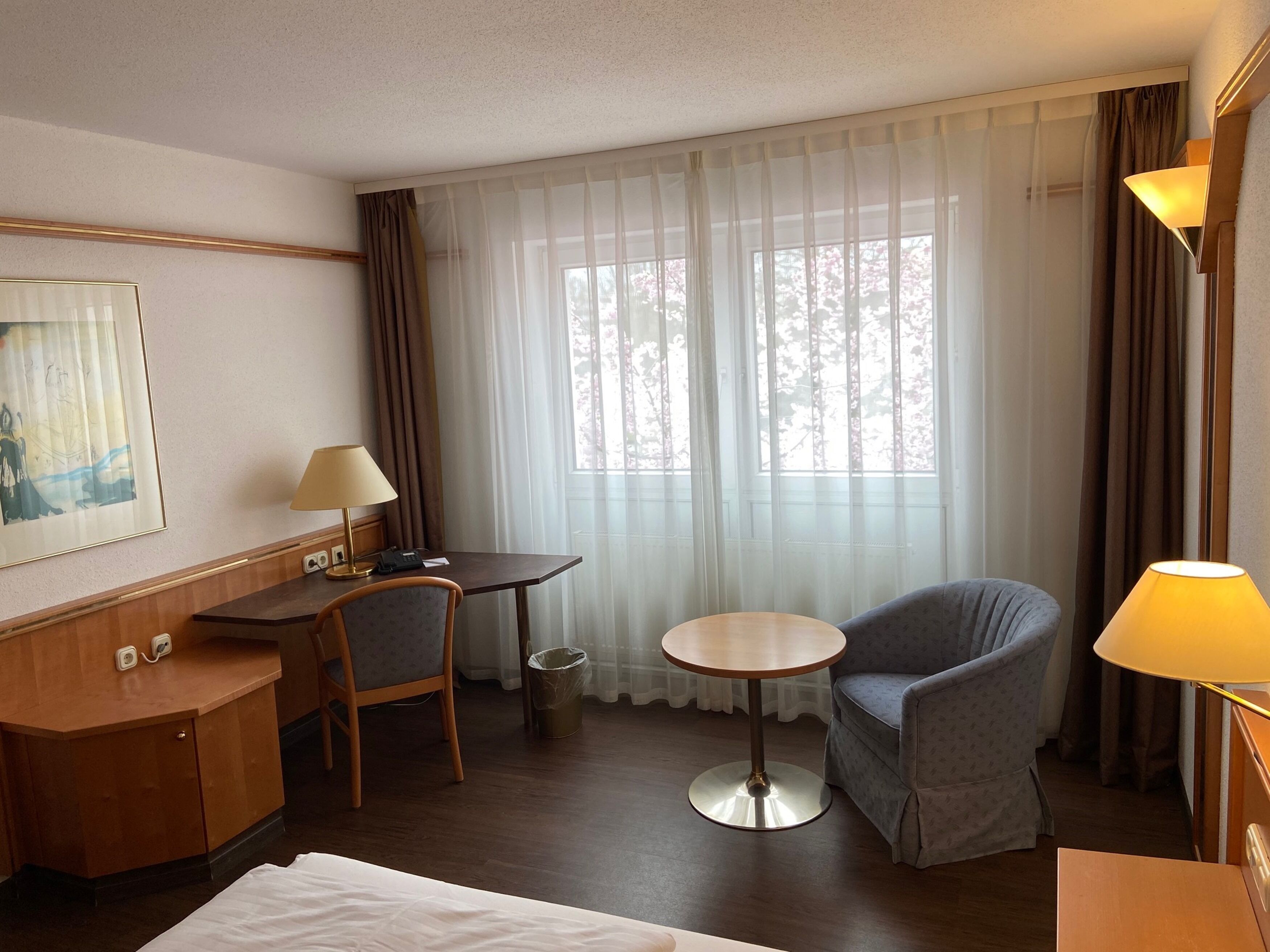 Une chambre d’hôtel avec un lit, un bureau avec une lampe, une chaise, une petite table et une fenêtre avec des rideaux.