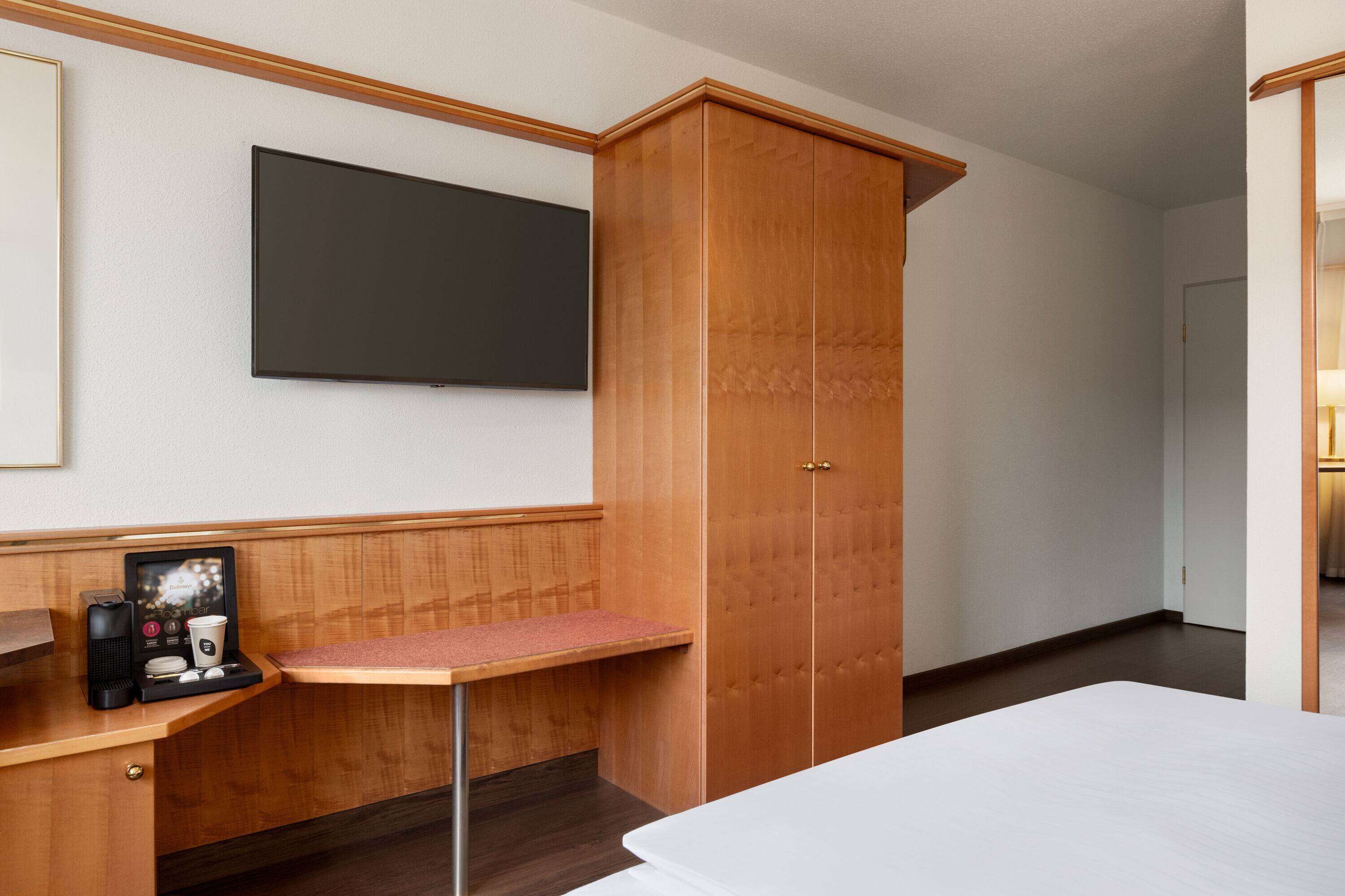 Une chambre d’hôtel comprenant un lit, une armoire en bois, un petit bureau avec une machine à café et un téléviseur à écran plat fixé au mur.