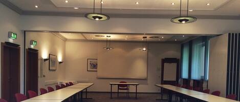 A conference room with long tables, red chairs, a whiteboard, and pendant lighting.