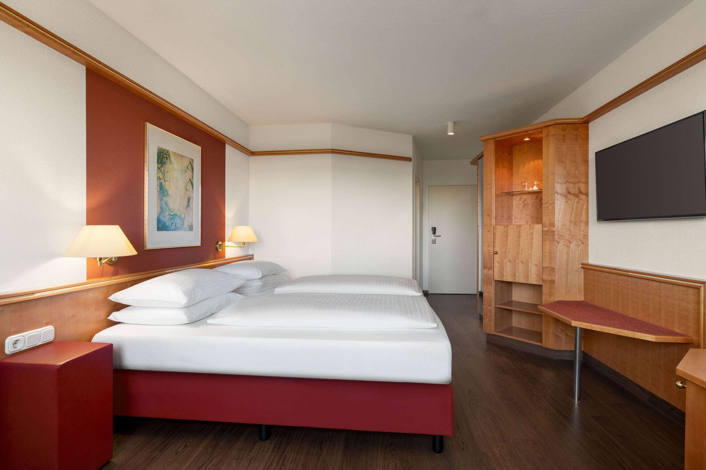 A hotel room with a bed, two bedside lamps, a TV mounted on the wall, and a wooden cabinet with a shelf.