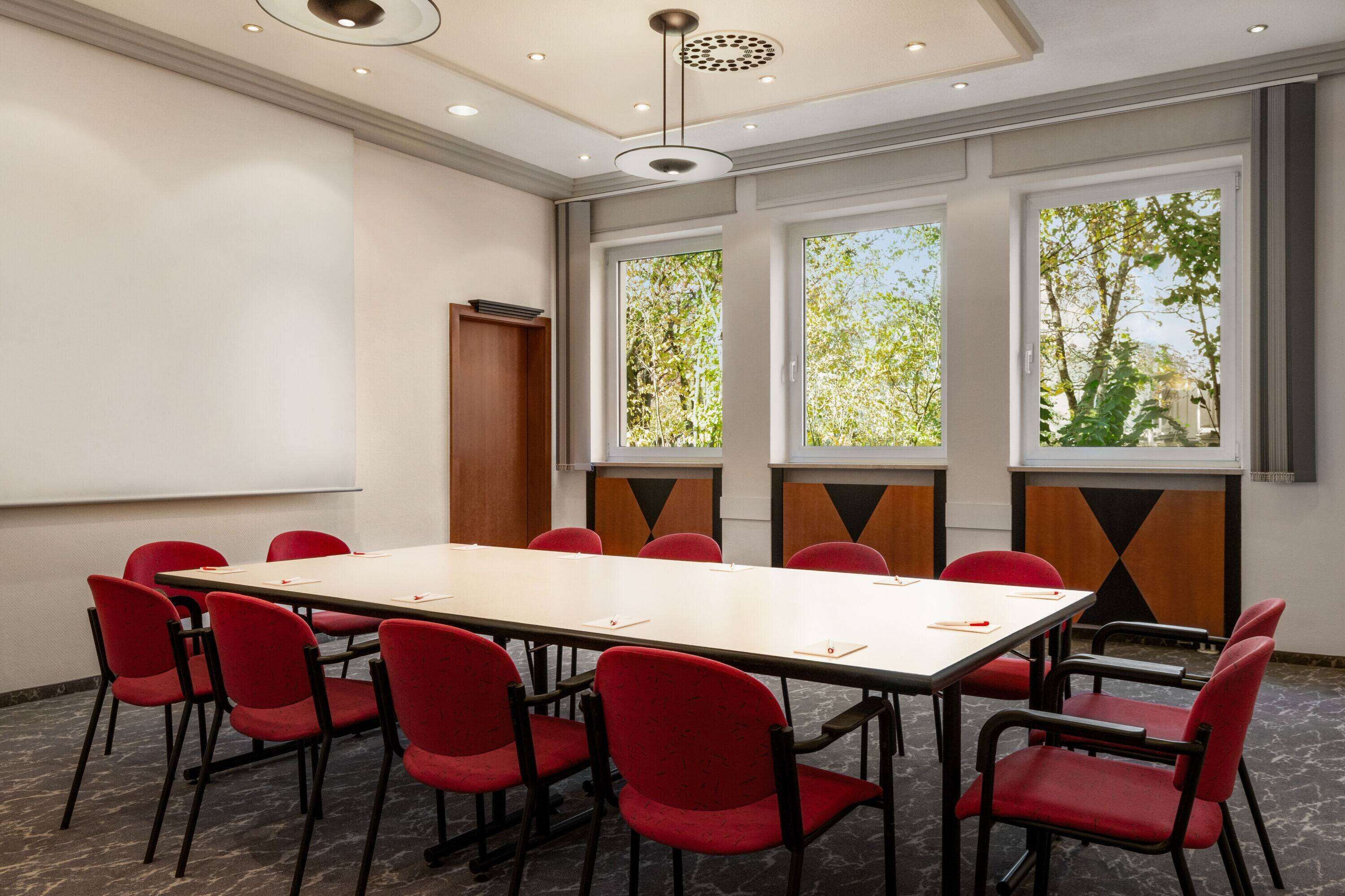 Ein Konferenzraum mit langem Tisch, roten Stühlen und großen Fenstern mit Blick auf Bäume.