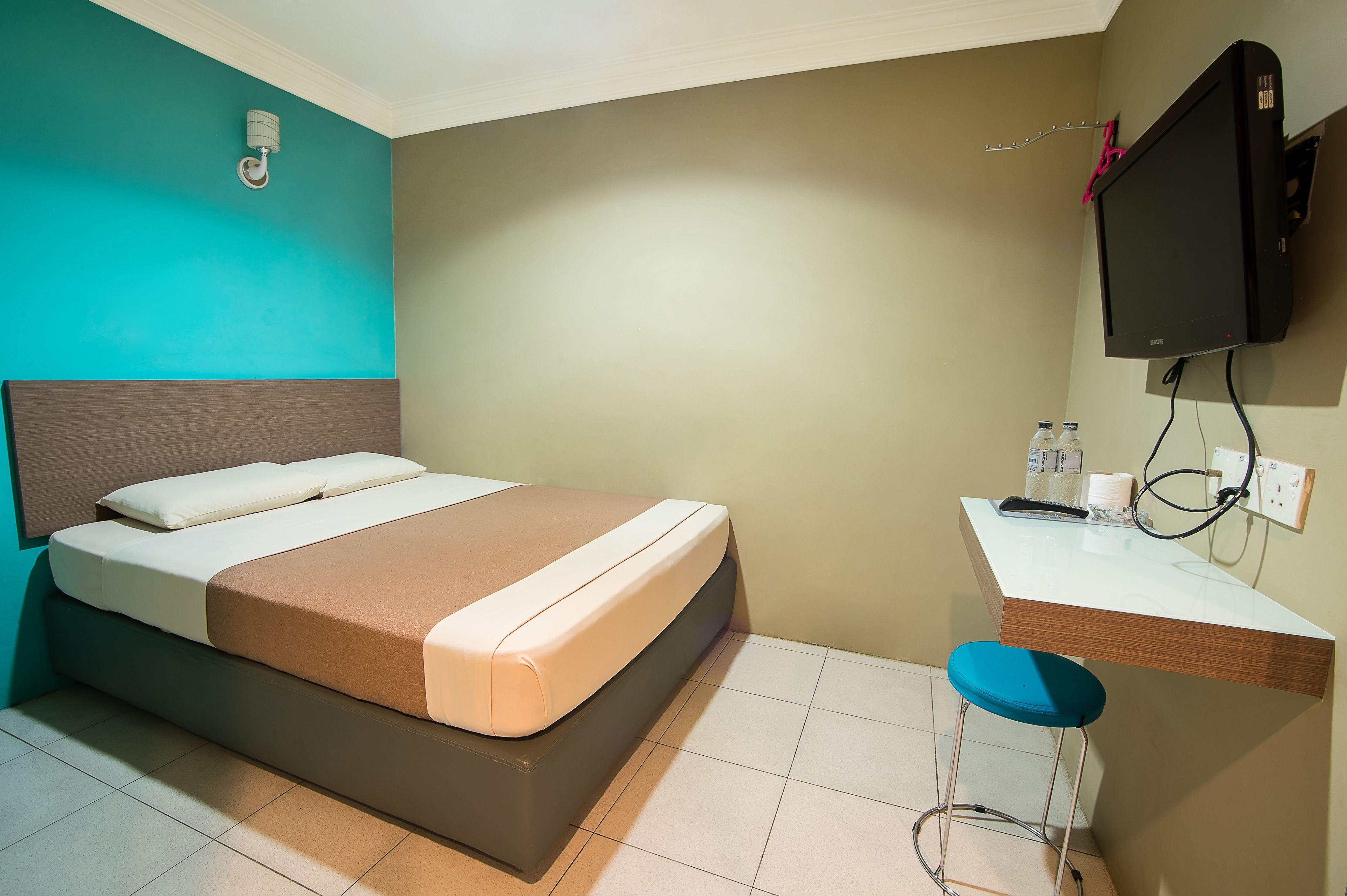 A hotel room with a bed, a desk with a chair, a TV, and a wall-mounted light fixture.