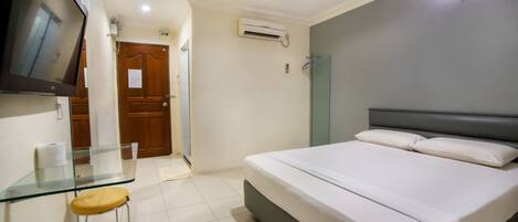 A bedroom with a bed, a glass table with a stool, a TV mounted on the wall, and an air conditioning unit.