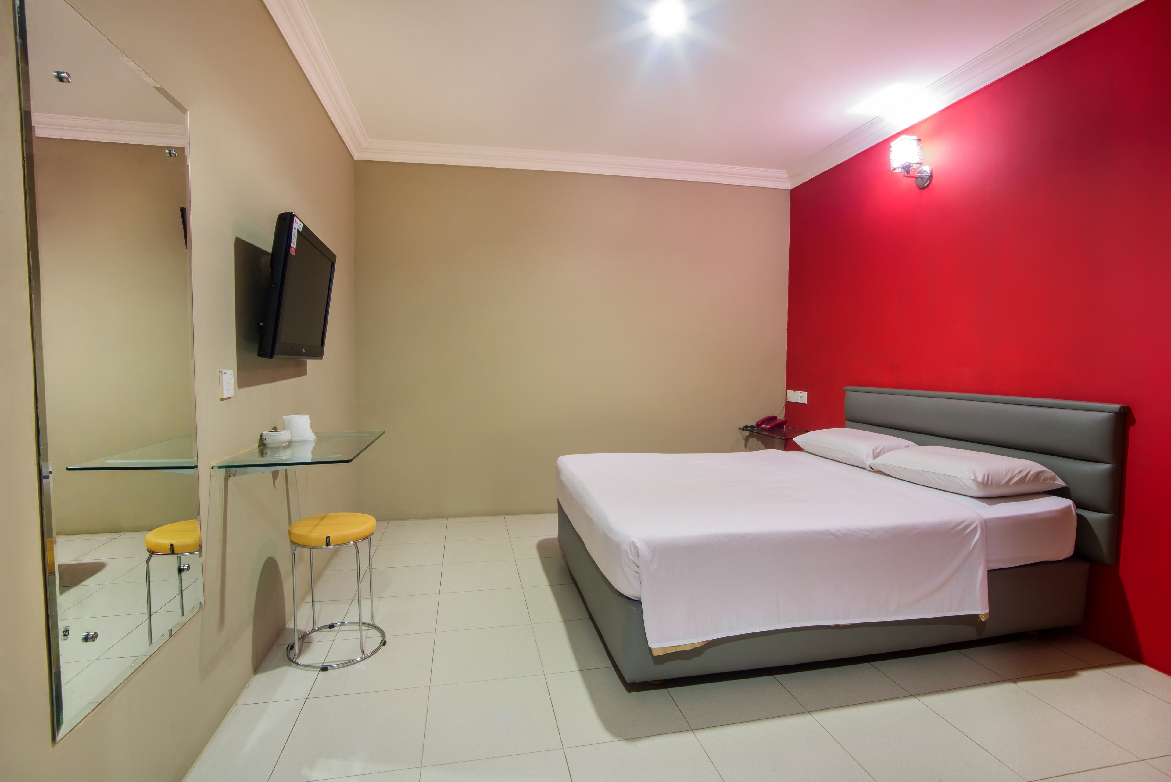 A bedroom with a bed, a small table with a chair, a mirror, and a television on the wall.