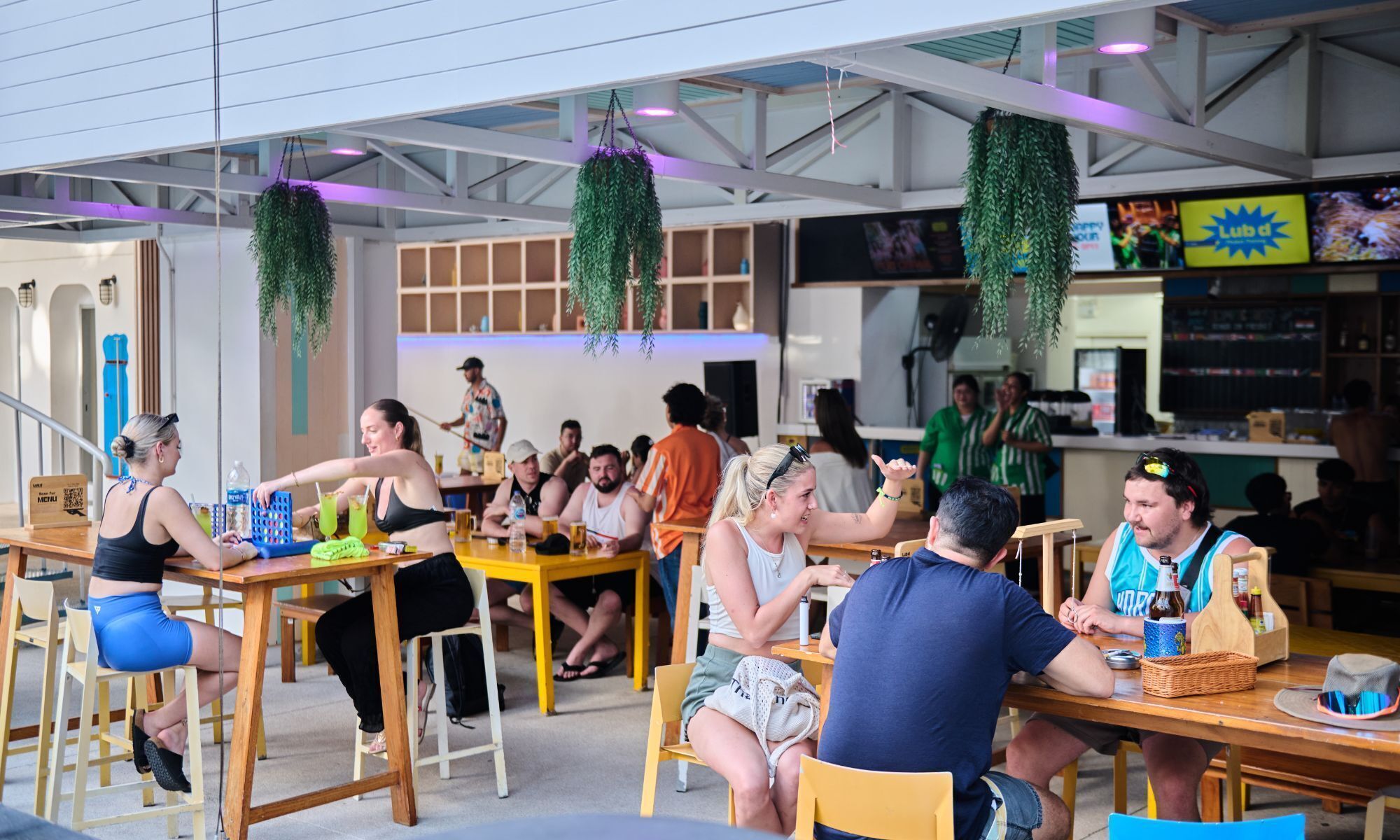 An indoor dining area with wooden tables and chairs, hanging plants, and a menu board.