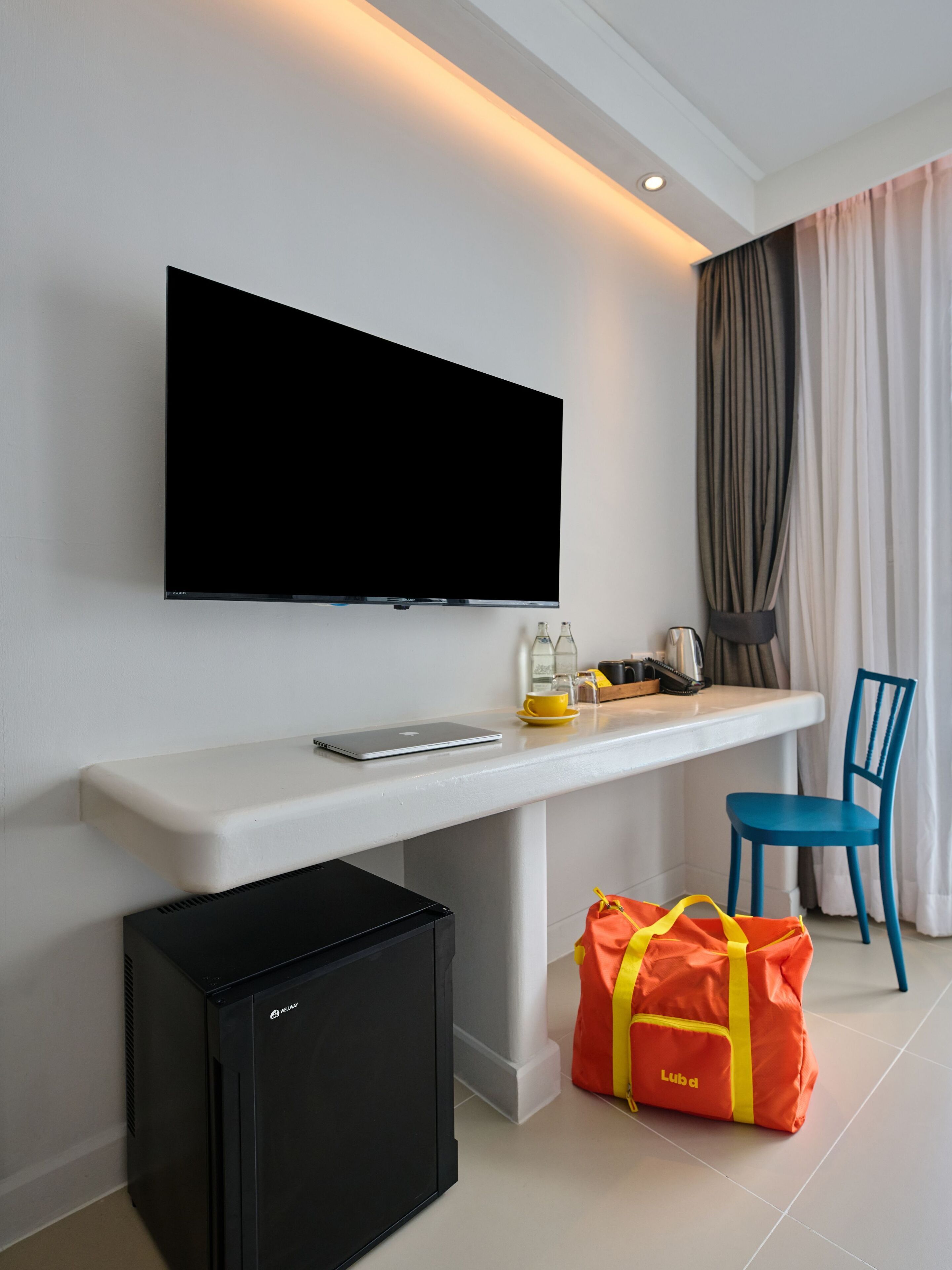A modern hotel room with a flat-screen TV mounted on the wall, a small black refrigerator, a desk with a laptop, a yellow cup, and a red bag labeled "Lubd".