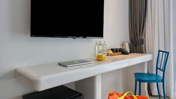 A modern hotel room with a flat-screen TV mounted on the wall, a small black refrigerator, a desk with a laptop, a yellow cup, and a red bag labeled "Lubd".