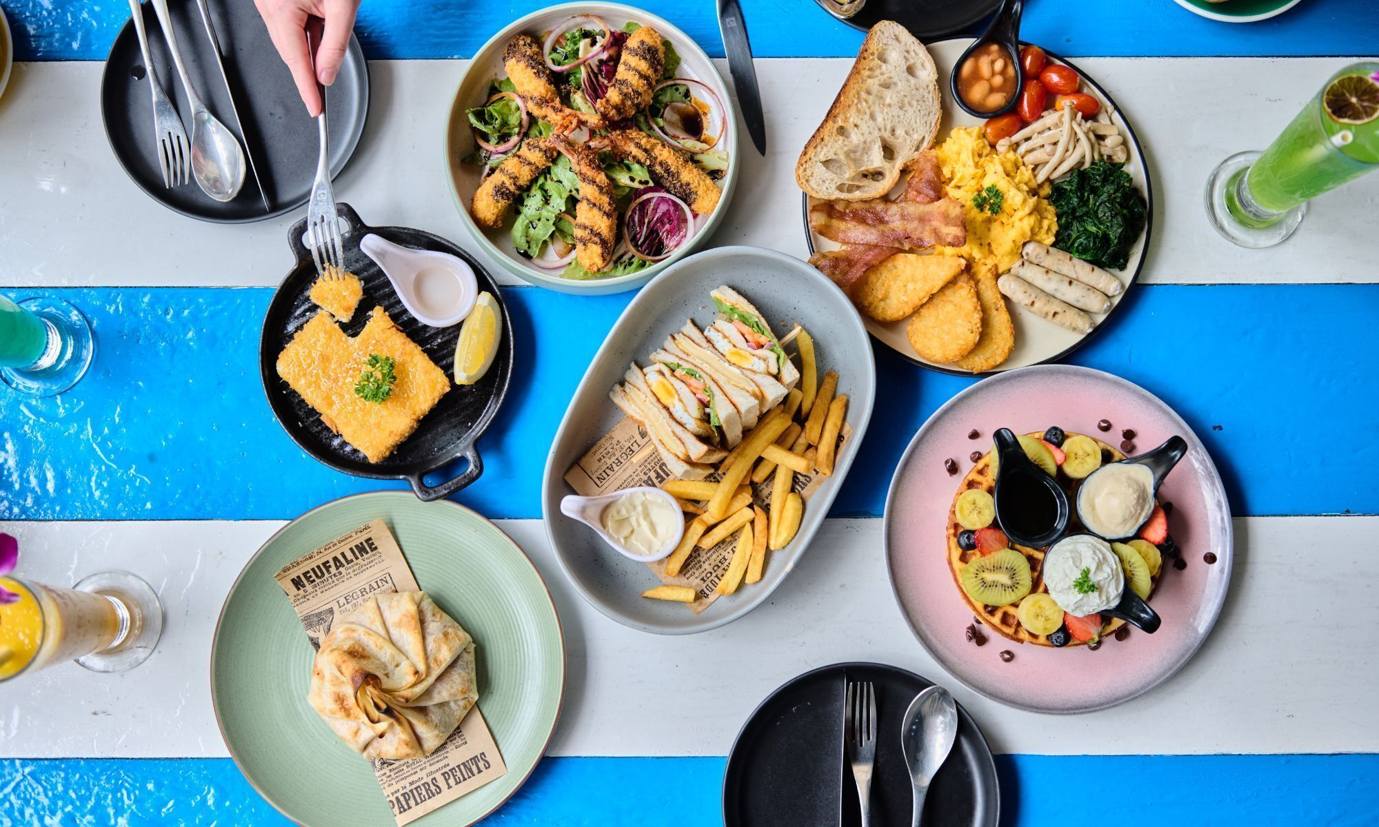 Une table garnie de différents plats pour le petit-déjeuner, notamment des œufs brouillés, des saucisses, des toasts et une assiette de crêpes et de fruits.
