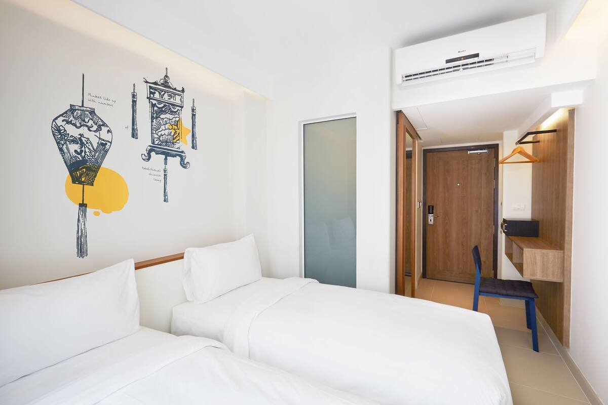A hotel room with two beds, a wall-mounted air conditioner, a door leading to a bathroom, and a small desk with a chair.