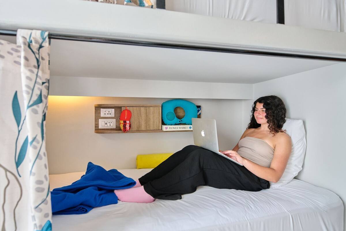 A bunk bed with a laptop on the lower bunk, a pillow, a blanket, and a wall-mounted shelf with a red object and a blue pillow.