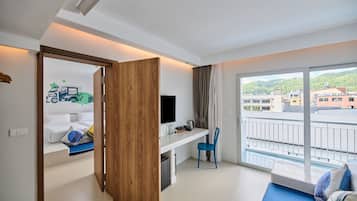 A hotel room with a bed, a desk with a TV, a chair, and a balcony with a view of buildings and hills.