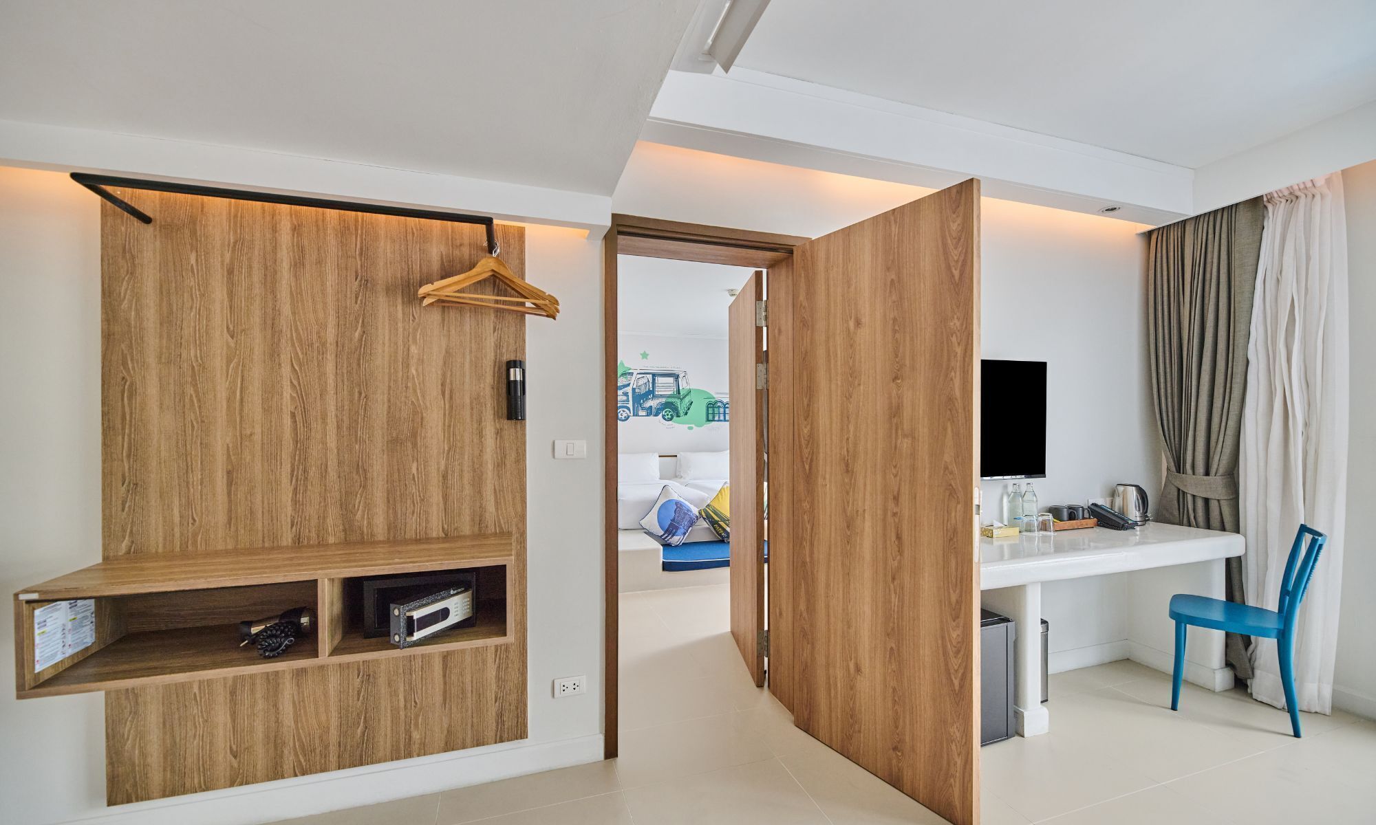 A modern hotel room with a wooden wardrobe, a desk with a chair, a TV, and a view of the outside.