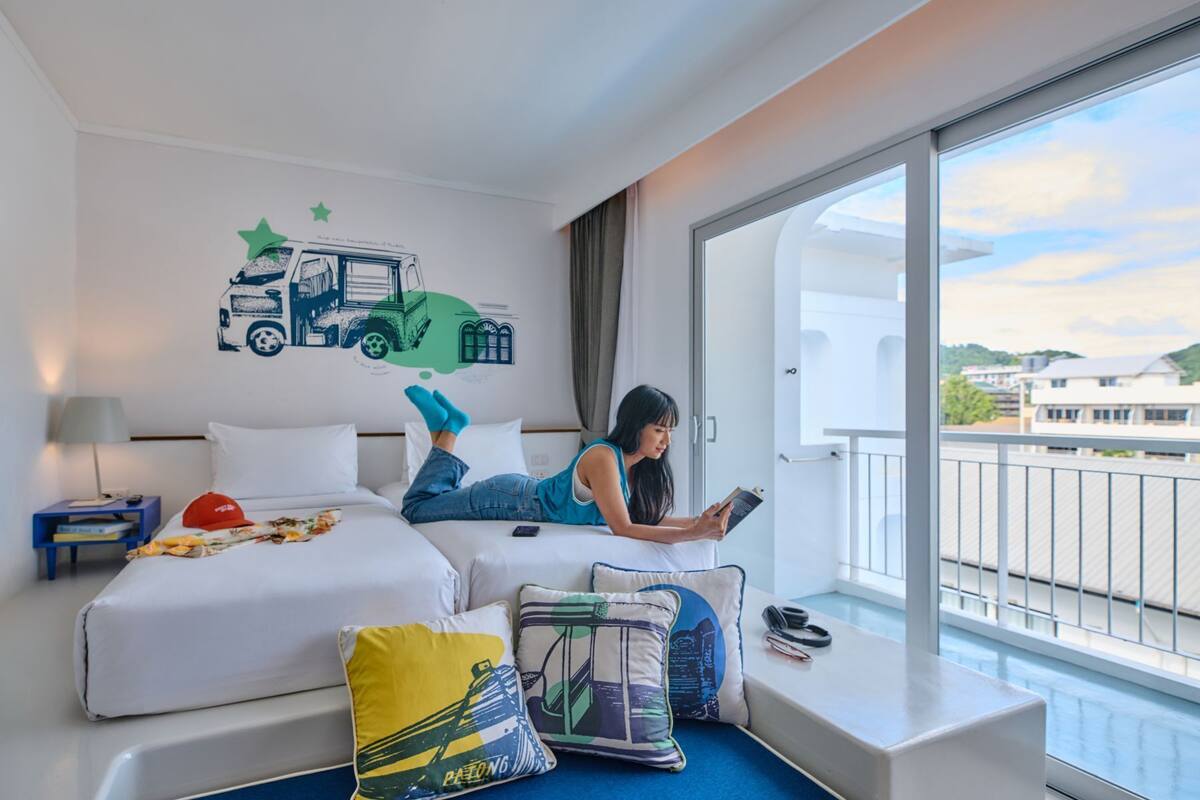 A hotel room with two beds, a large window with a view of buildings, and a wall decal of a truck.