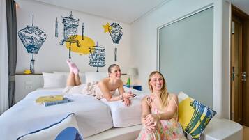 Hotellihuone, jossa on sänky, kaksi ihmistä makaa siinä, yöpöydällä on lamppu ja seinällä on taideteos.