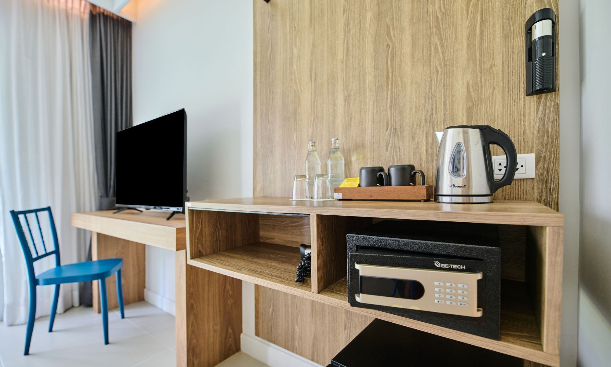A compact hotel room with a wooden desk, a television, a microwave, a kettle, and a chair.