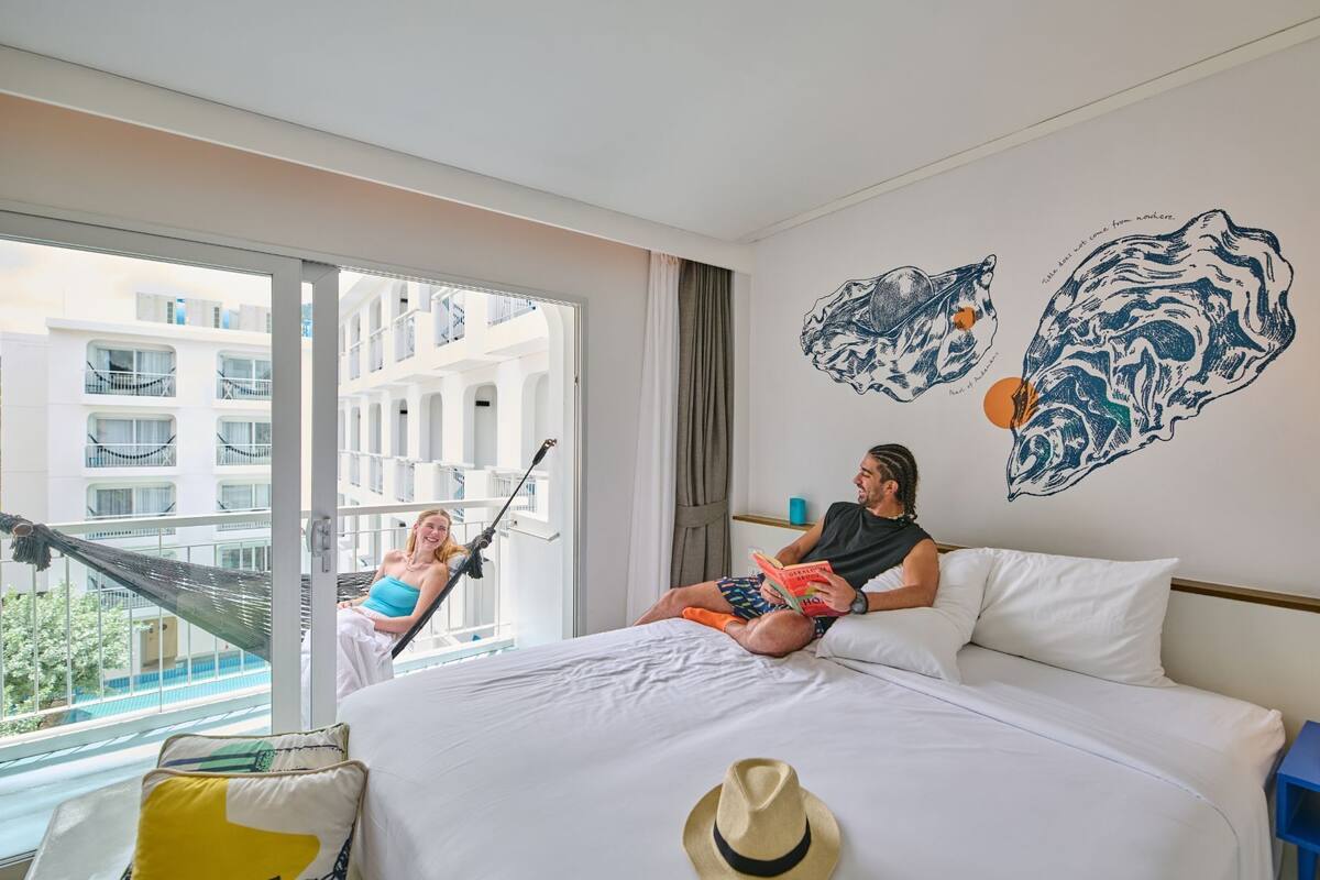 A hotel room with a large bed, a hammock, and a view of a building through a large window.