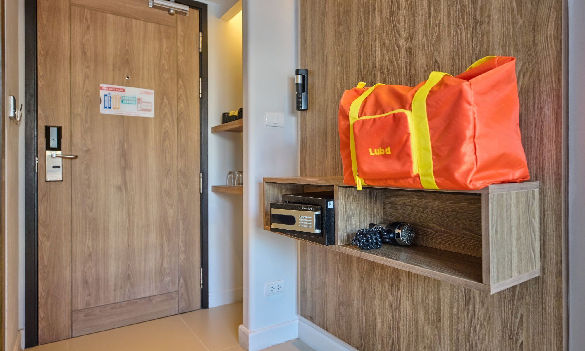 A hotel room with a wooden door, a shelf with a Lubb bag, a thermostat, and a small decorative item.