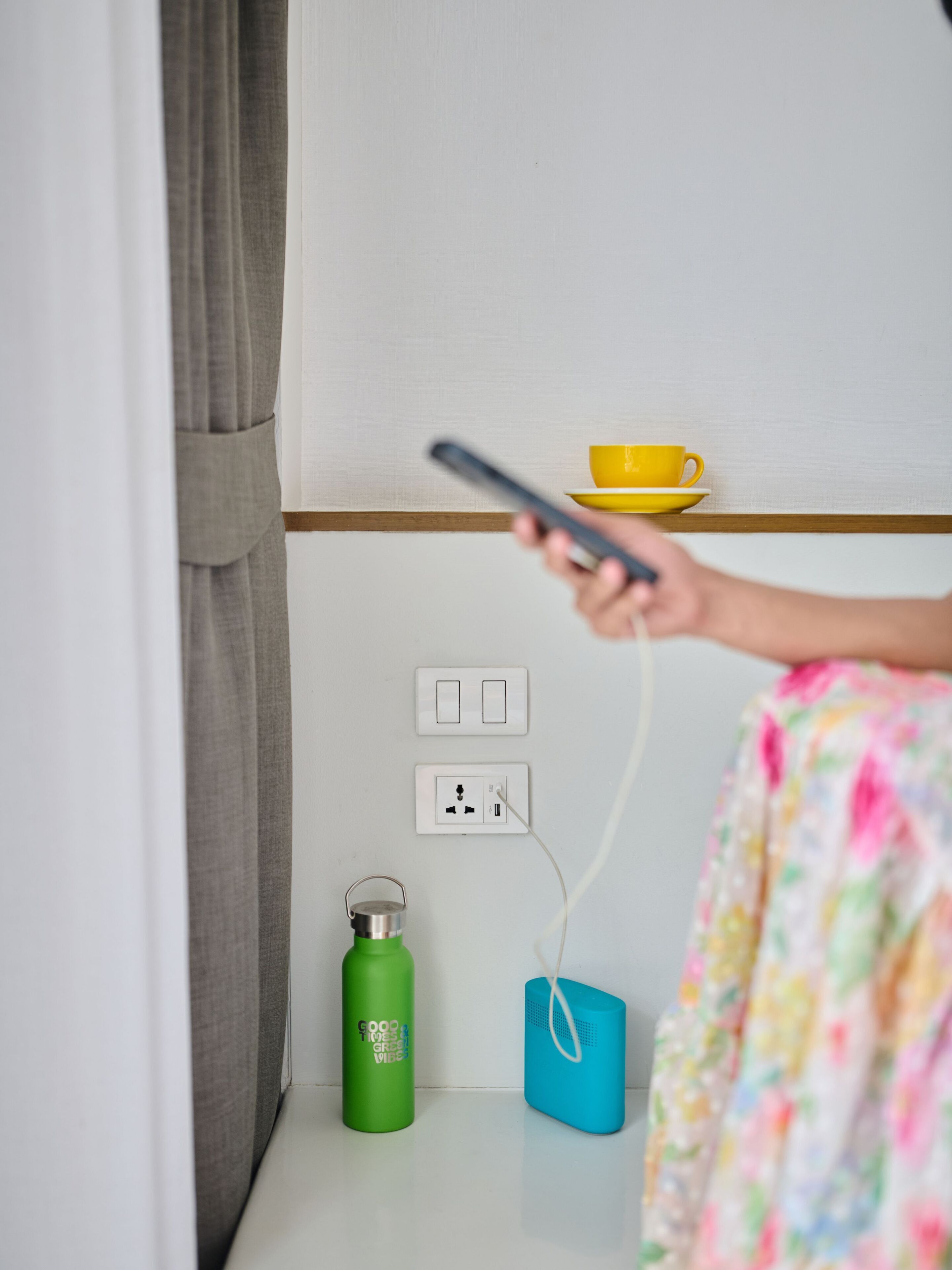 A room with a bed, a bedside table, a yellow cup, a green water bottle, a blue speaker, and a power outlet.