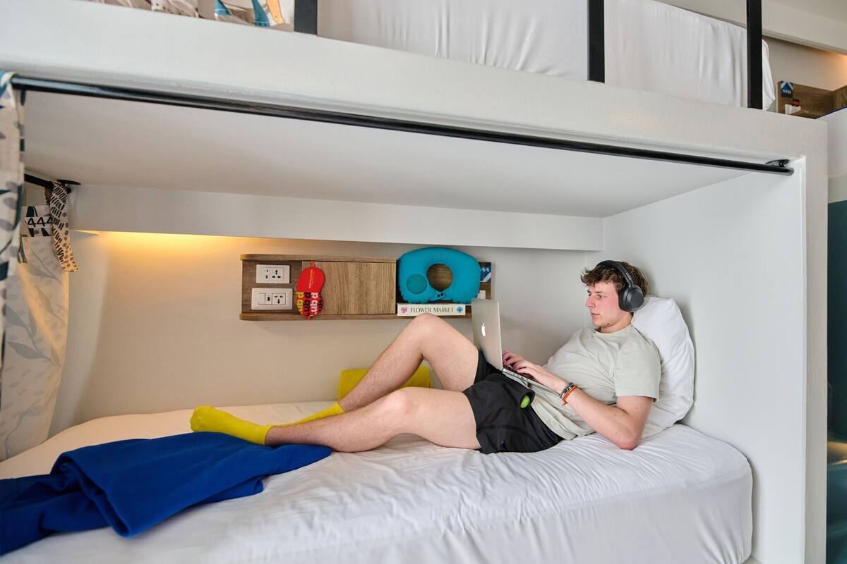 A bunk bed with a person using a laptop. There is a shelf with a pillow, a small rug, and a sign that says "Flower Market".