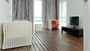 A room with a wooden floor, a white wicker sofa with a red cushion, a black TV stand, and a large window with grey curtains. - Laman Pesona Resort & Spa (Raub)