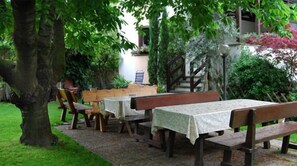 An outdoor seating area with wooden benches and tables, surrounded by lush greenery and a stone pathway. - Garni Meinrad (Ora)