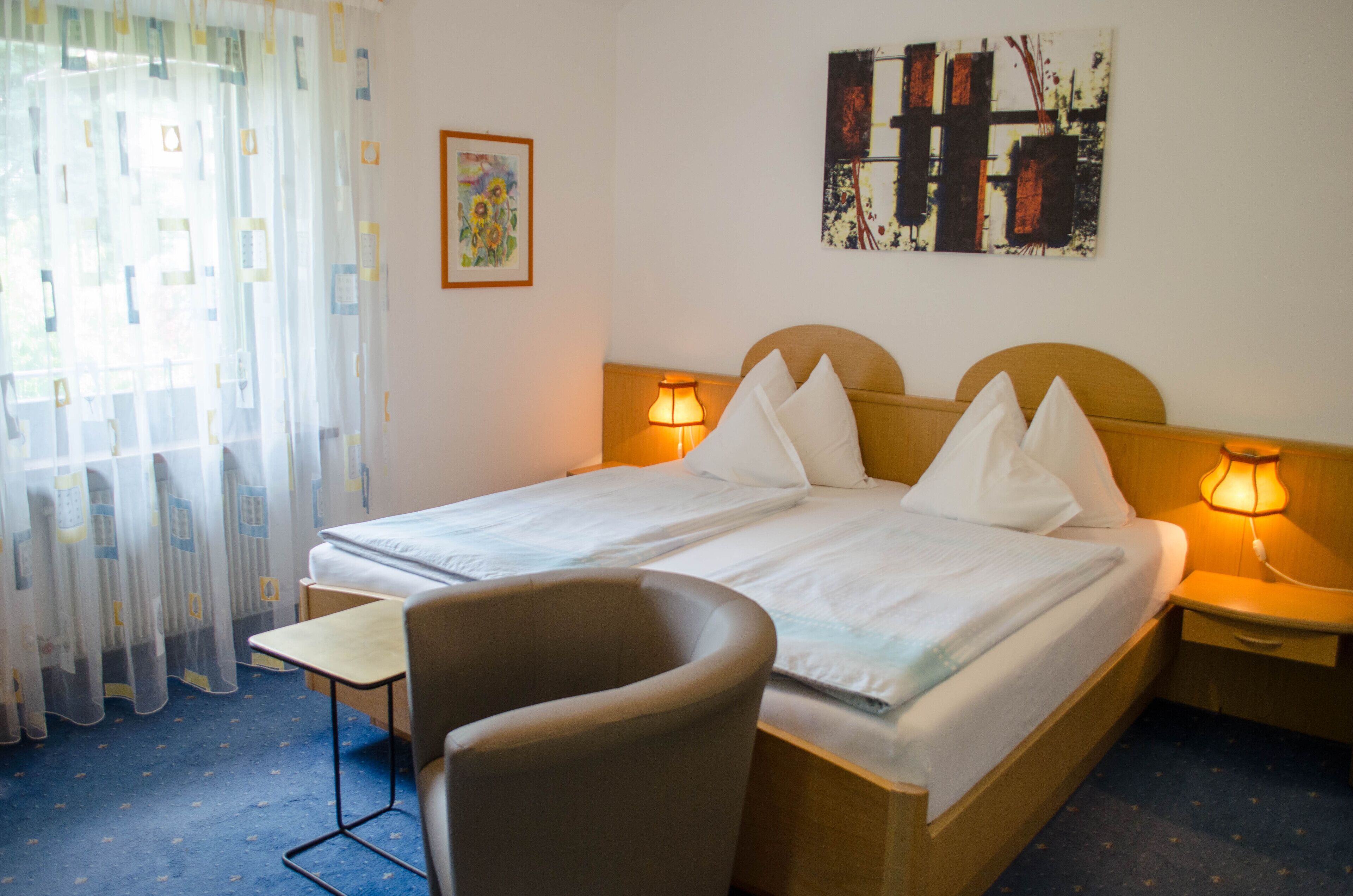 Habitación de hotel con cama, lámparas de noche, una silla, una mesita y obras de arte en las paredes.