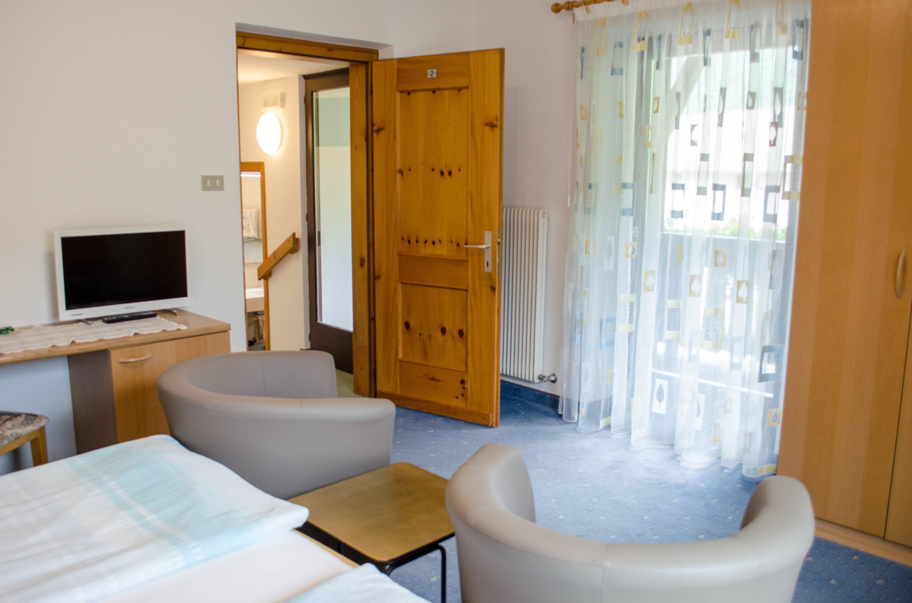 Een kamer met een bed, een bureau met een computer, twee fauteuils, een houten deur en een raam met gordijnen.