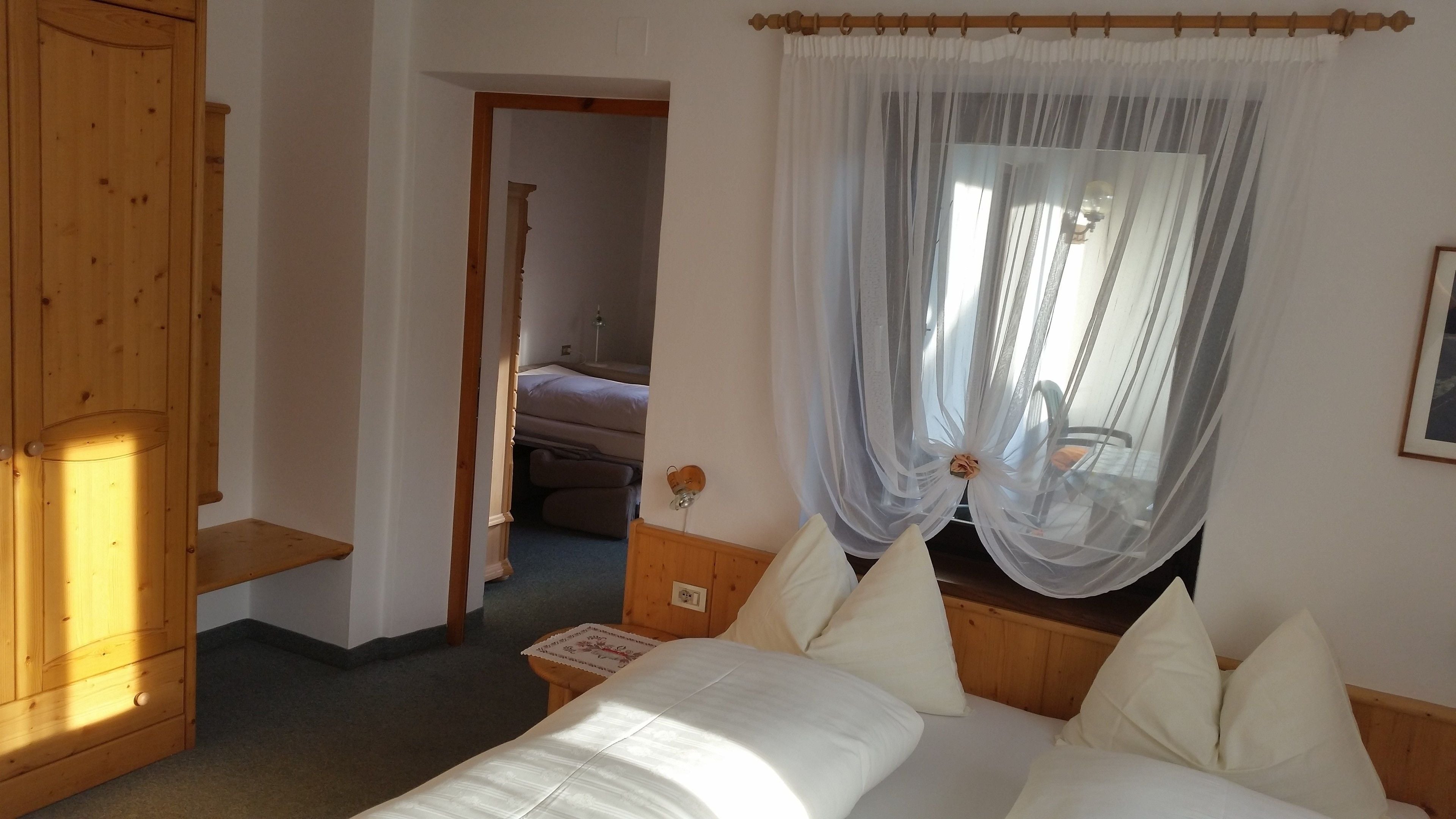 A bedroom with a wooden wardrobe, a bed with white bedding, and a window with sheer curtains.