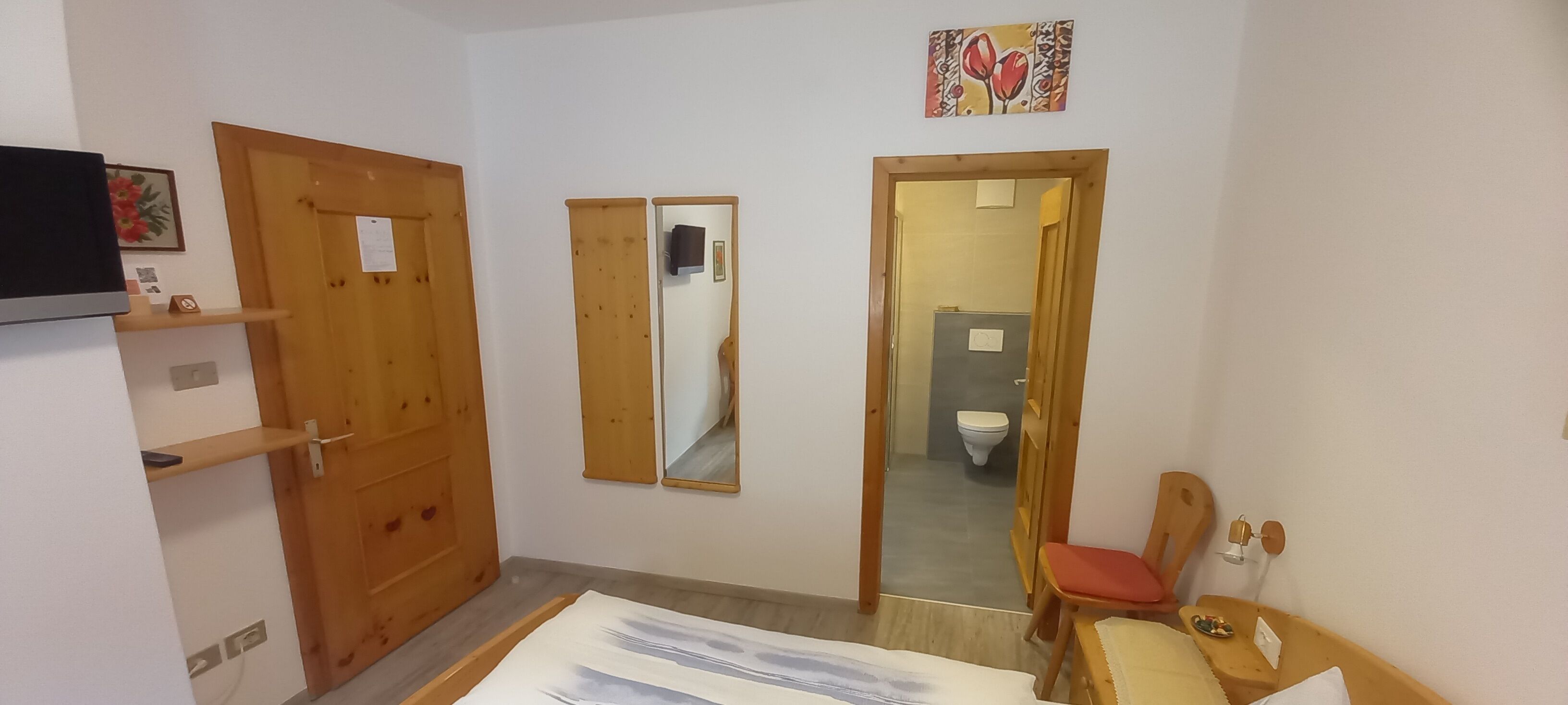 A room with a wooden door, a mirror, a bed, a chair, and a small table.