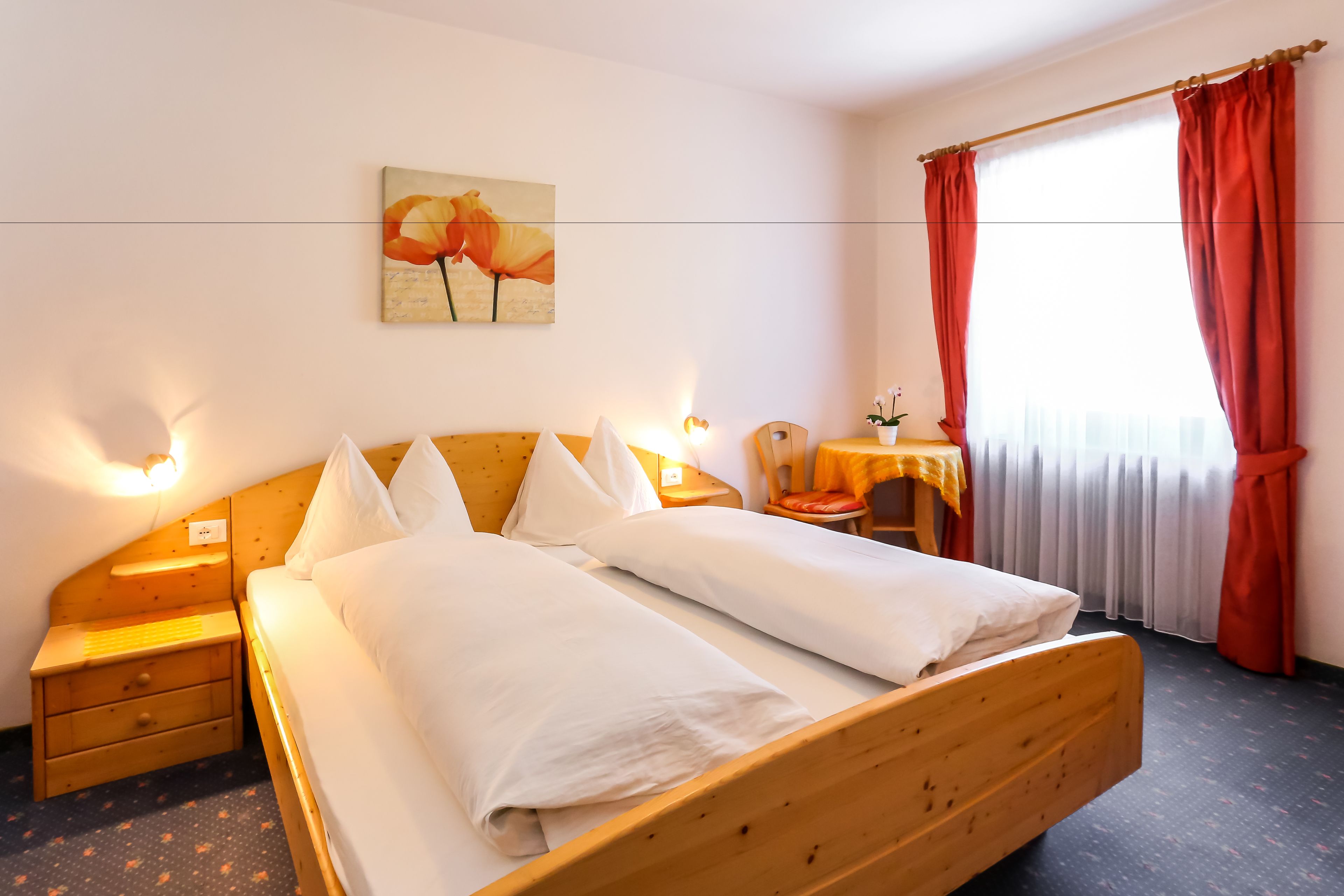 A hotel room with a wooden bed, two bedside tables, a nightstand, a small table with a chair, and a window with red curtains.