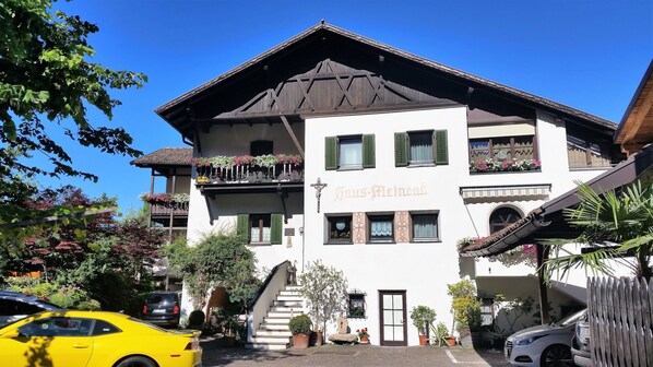 A multi-story building with a traditional Alpine architectural style, featuring balconies with flower boxes, a staircase leading to the entrance, and a sign reading "Sous-Mont." - Garni Meinrad (Ora)
