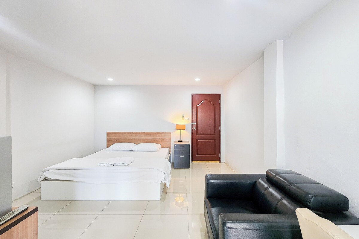 Une chambre d’hôtel moderne, équipée d’un lit, d’un bureau avec une lampe, d’un canapé en cuir noir et d’une porte en bois.