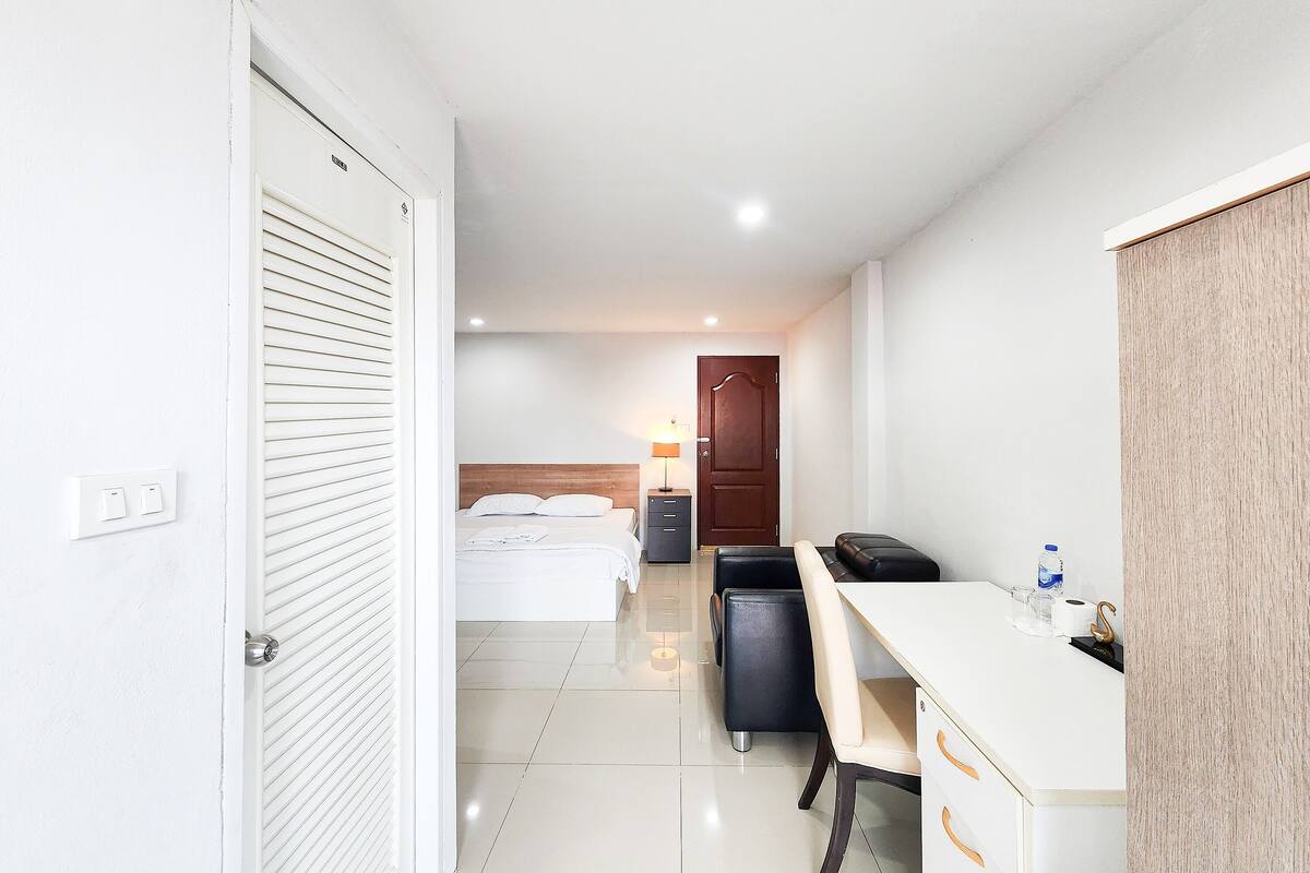 A hotel room with a bed, desk, chair, and a small table with a lamp and a bottle.