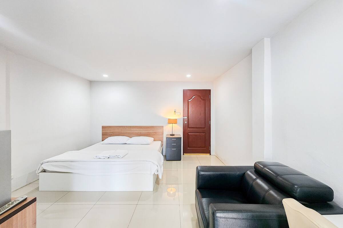 A modern hotel room with a bed, a desk with a lamp, a black leather sofa, and a wooden door.